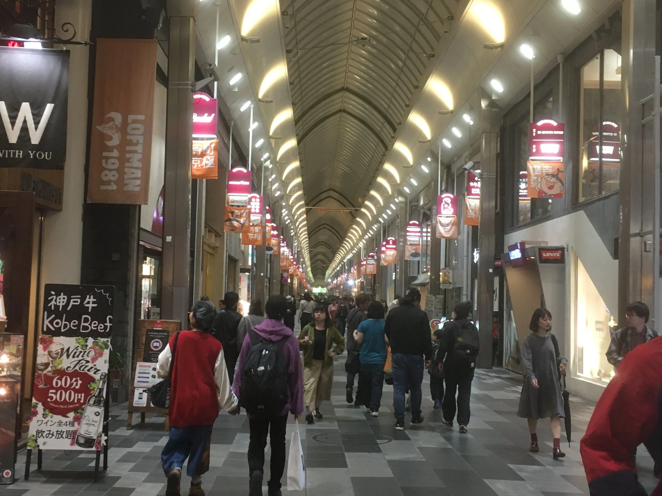 People walking through a covered shopping arcade with bright lights and storefronts on both sides, some holding shopping bags, and signs advertising food and wine offers.
