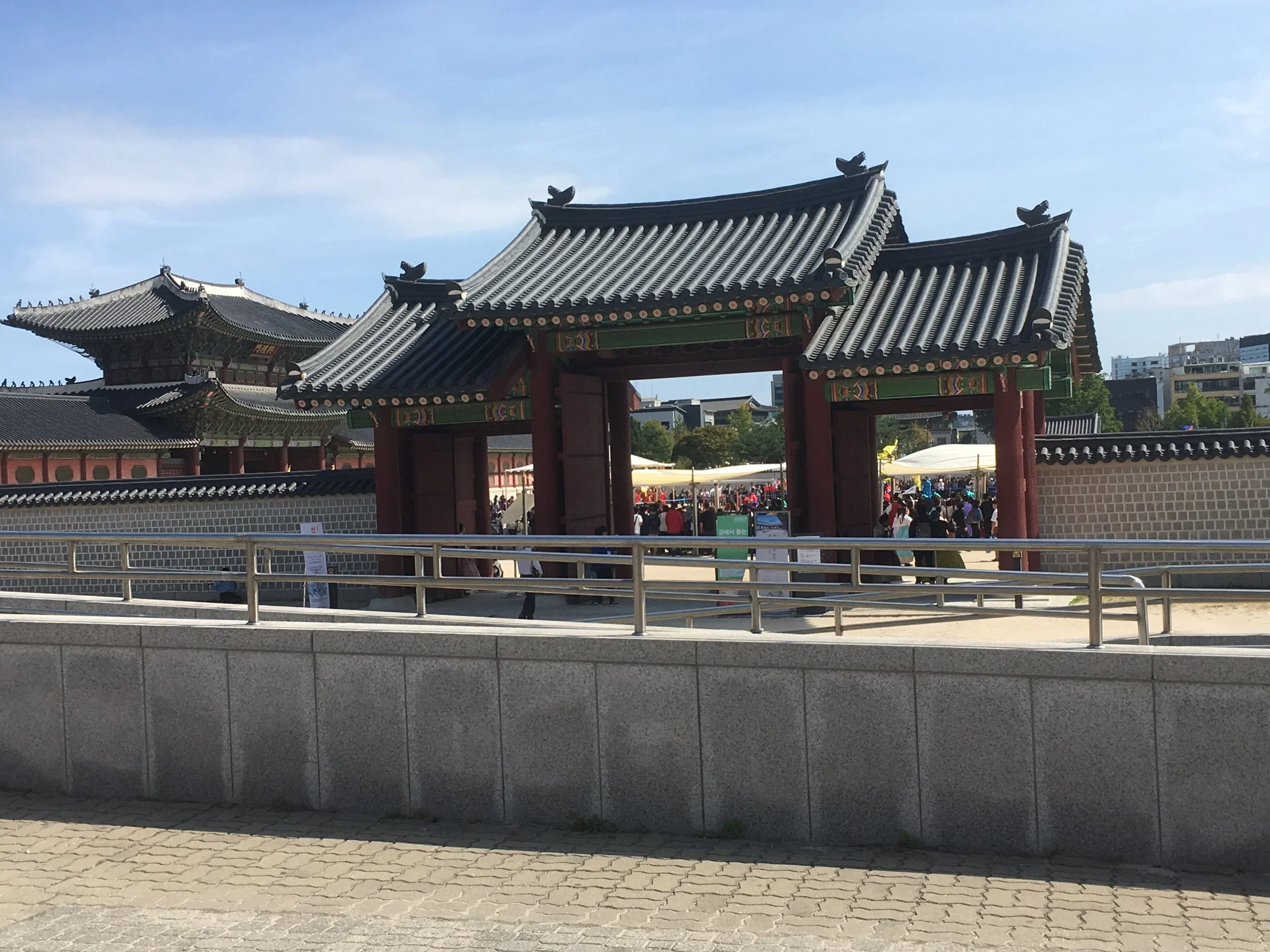Traditional Korean palace entrance with ornate tiled roofs and a bustling courtyard behind, sunny sky above.