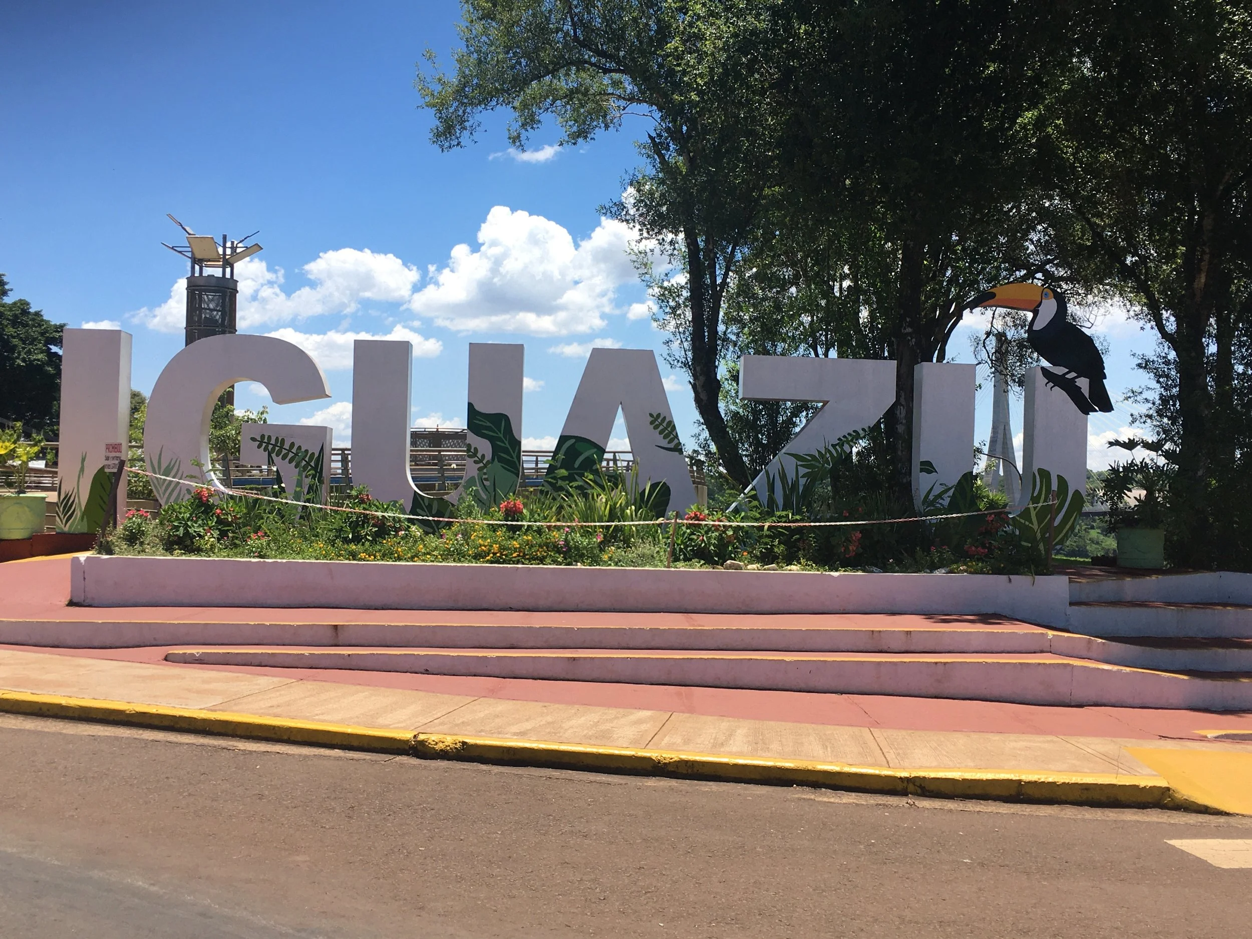 Large white letters spelling 'CUAZI' with painted green leaves, a toucan perched on the letter 'I', a garden with flowers in front, steps, and a clear blue sky with clouds in the background.