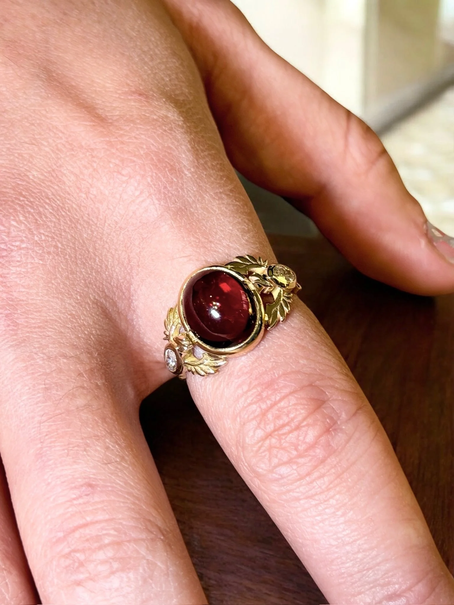 Close-up of a hand wearing a gold ring with a large oval red gemstone in the center, surrounded by small clear gemstones and decorative gold leaves.