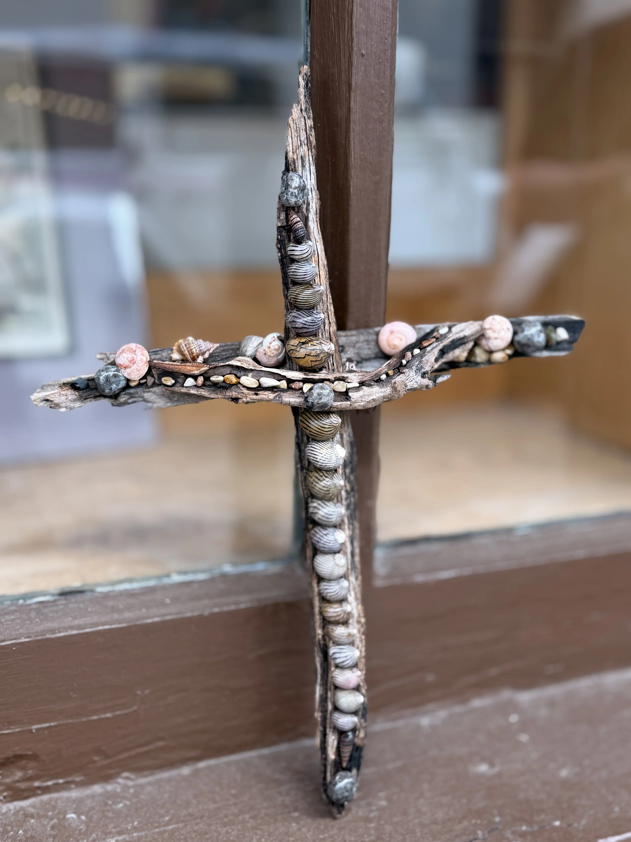 A cross made of weathered wood, decorated with small seashells, pebbles, and seed beads, mounted outdoors against a wooden and glass background.