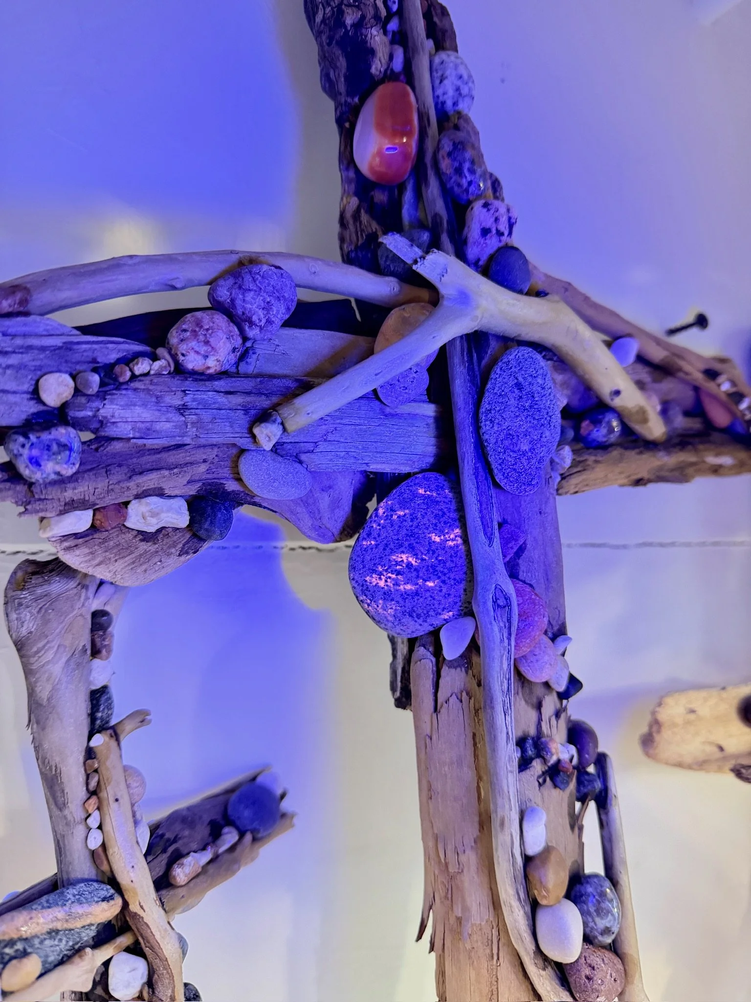 Close-up of a rustic cross made of driftwood and decorated with various small rocks and pebbles, illuminated with blue light.