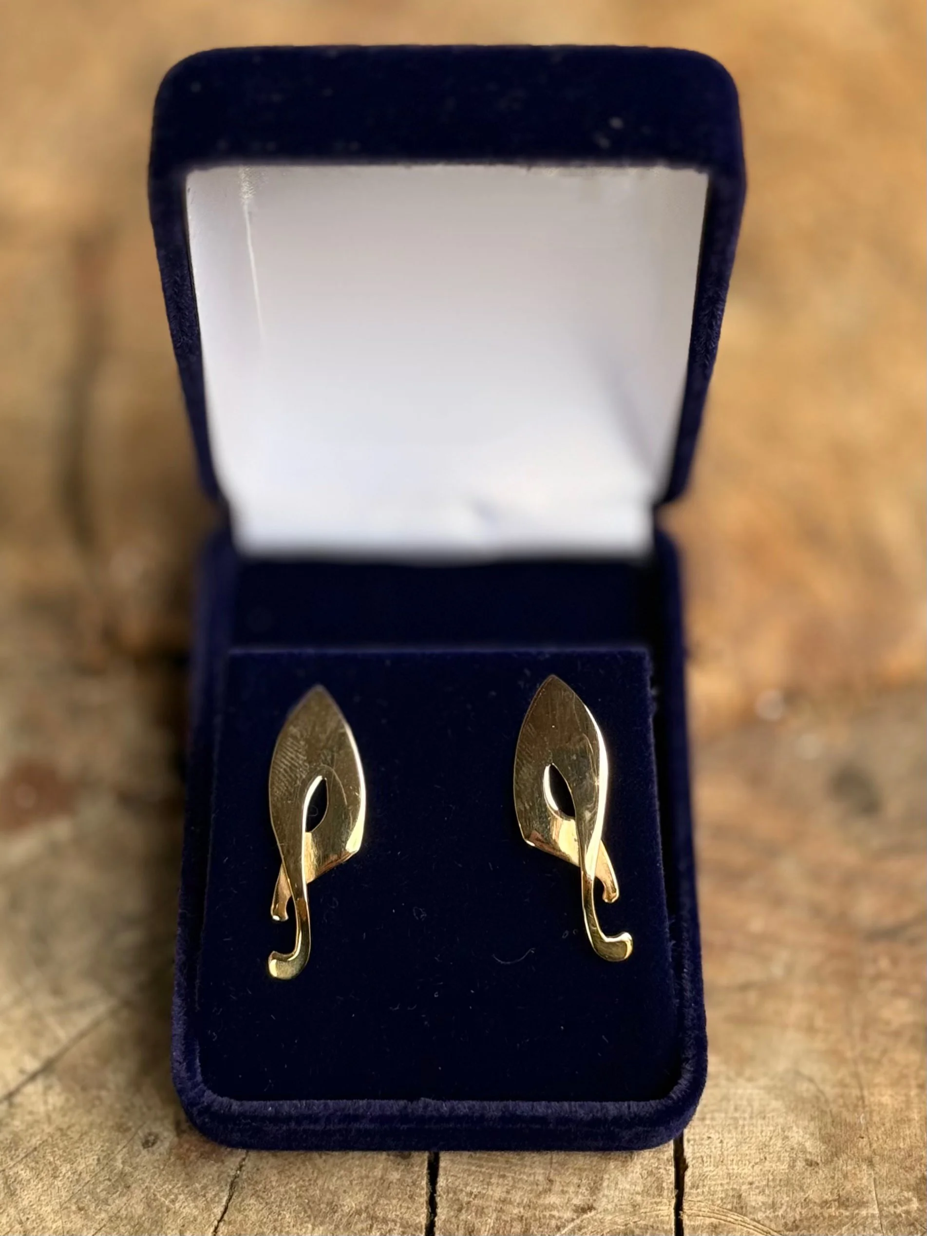 Gold earrings shaped like leaves displayed in a dark blue jewelry box with a white interior, placed on a wooden surface.