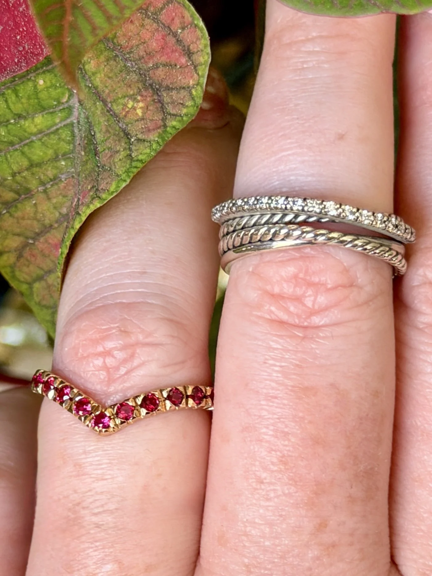 Close-up of a hand with three silver rings, one with small diamonds, another with a twisted design, and the third a plain band, on the middle finger, next to a leaf with green and reddish coloration.