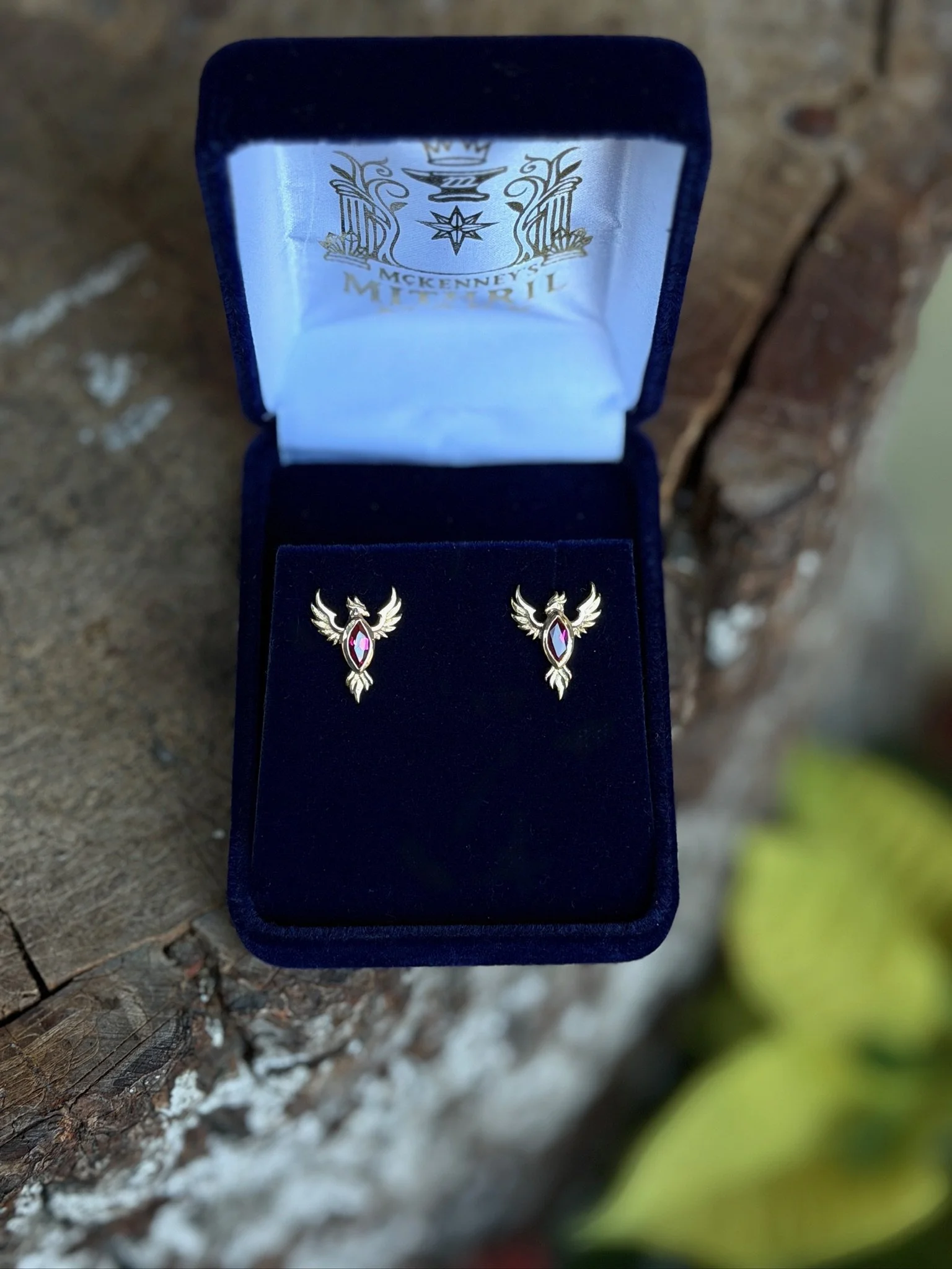 A pair of gold earrings with a phoenix design and pink stones inside a blue jewelry box with a printed interior.