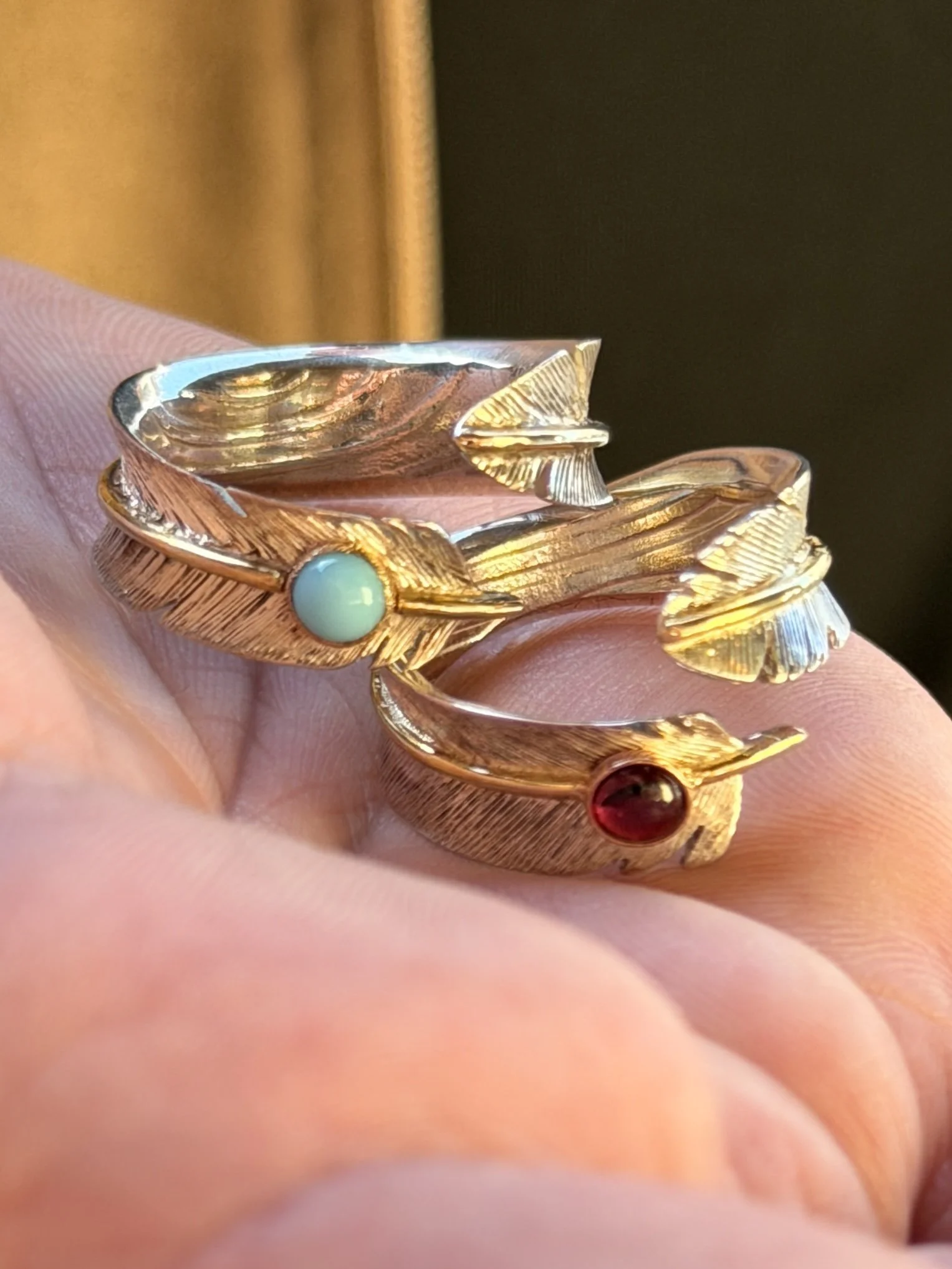 Close-up of a gold feather ring with a light blue gemstone and a gold feather ring with a red gemstone, held in a person's hand.