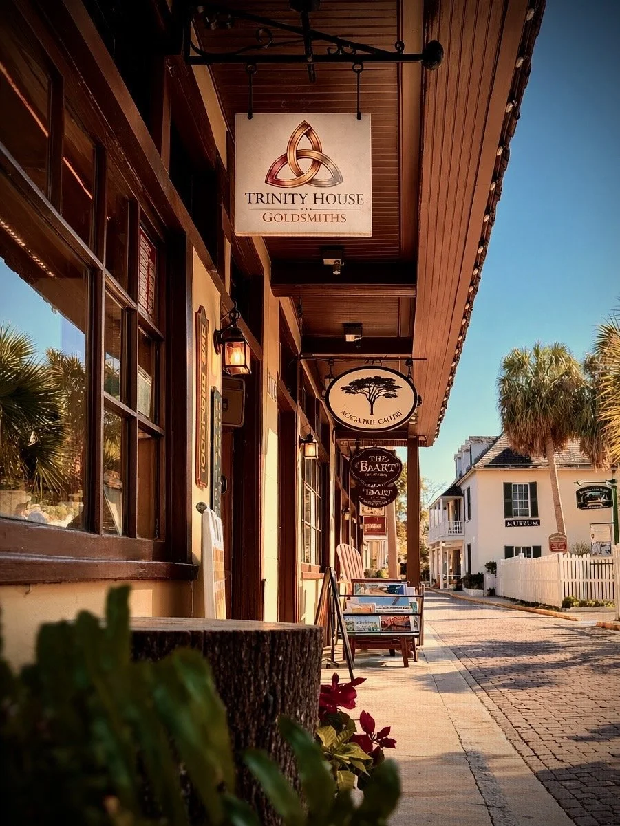 Street view in a small town with shops and palm trees, showing signs for Trinity House Goldsmiths, Acorn Tree Gallery, and The Baart, with outdoor chairs and a white picket fence.