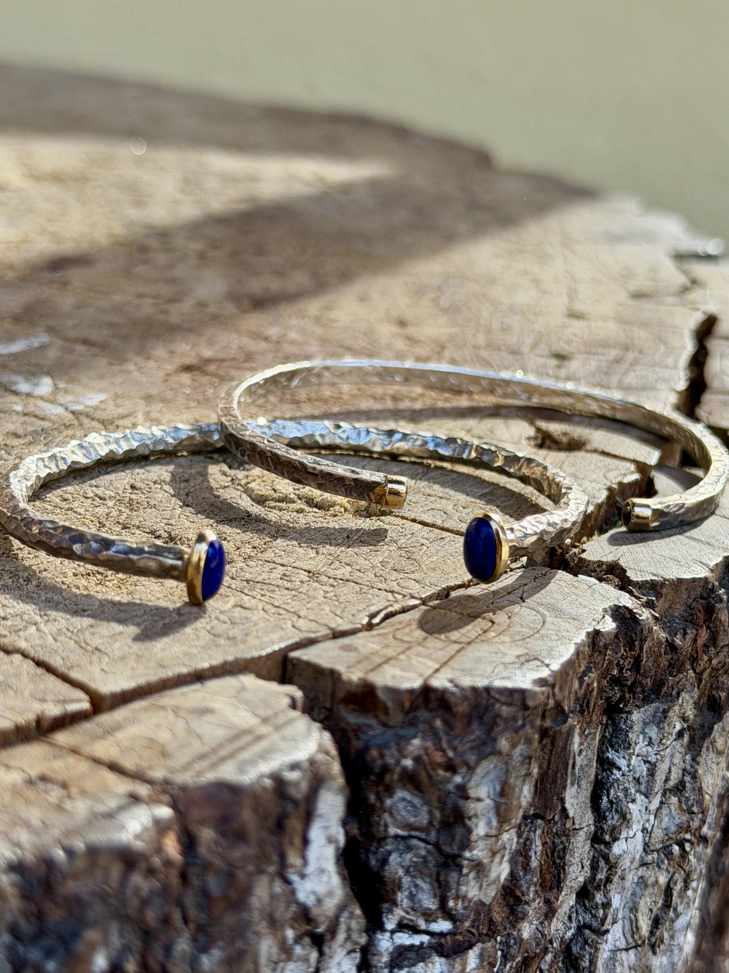Two hammered silver cuff bracelets with blue oval stones resting on a textured wooden surface.