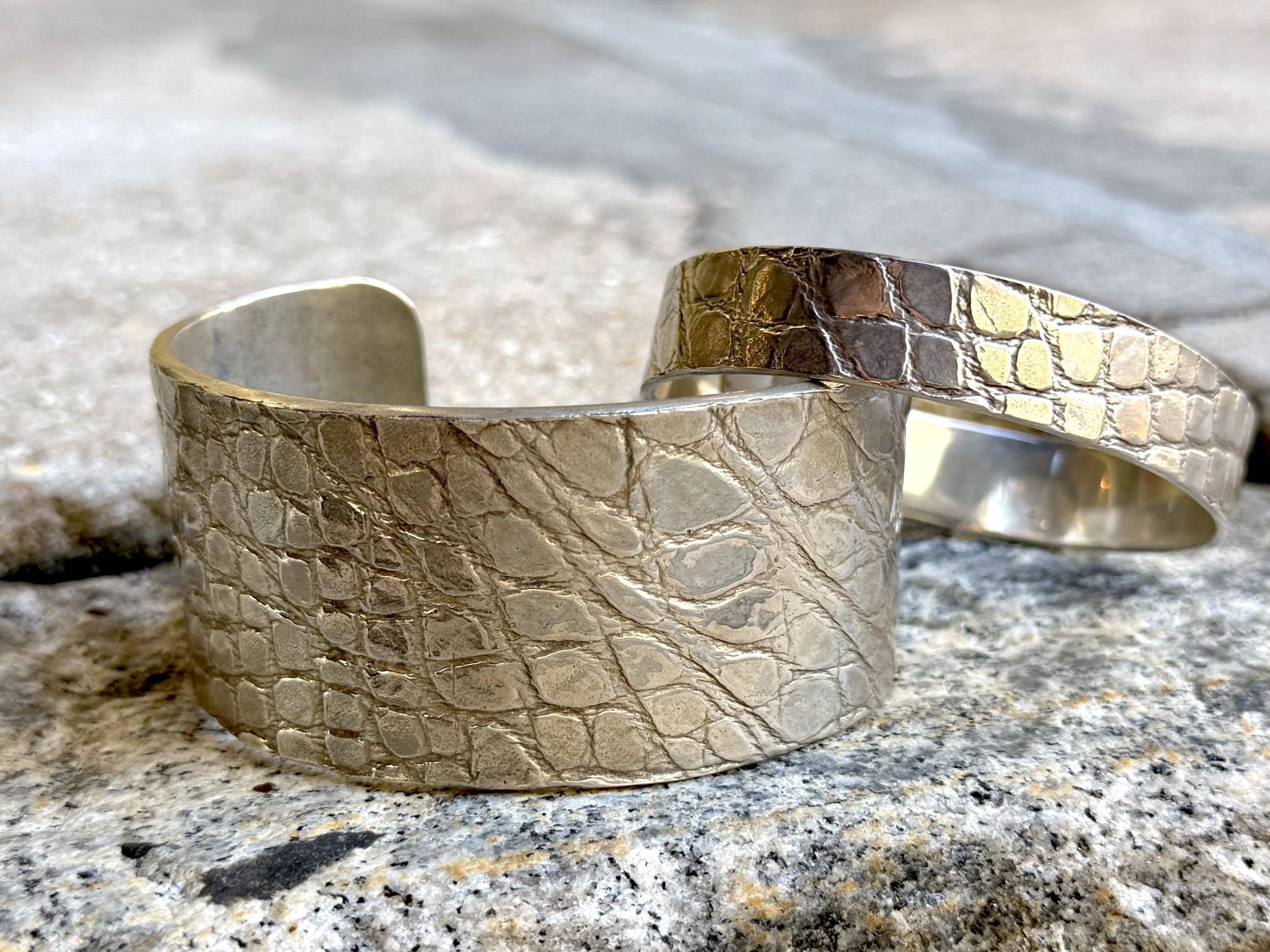 Two wide silver cuff bracelets with textured, scaled patterns resting on a rough stone surface.