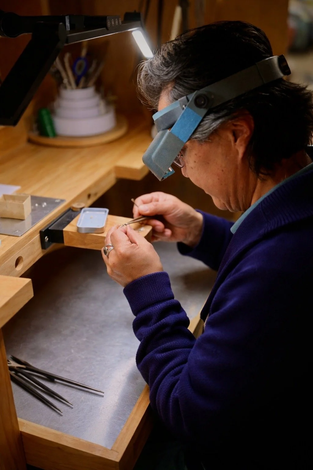 A person wearing a magnifying visor and glasses working carefully on a small object at a wooden workbench in a workshop, with tools and supplies in the background.