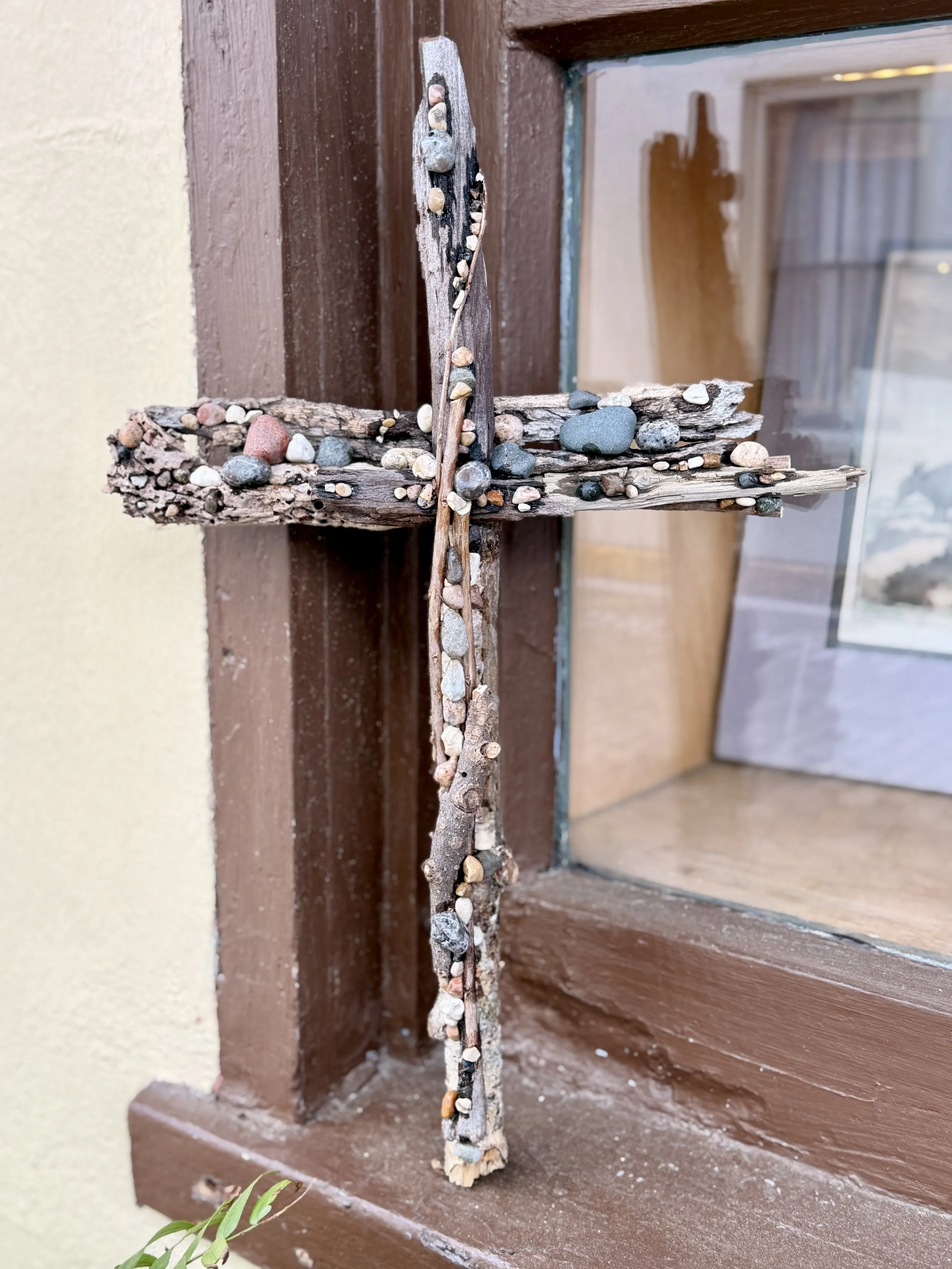 A cross made of small stones and driftwood affixed to a window frame.