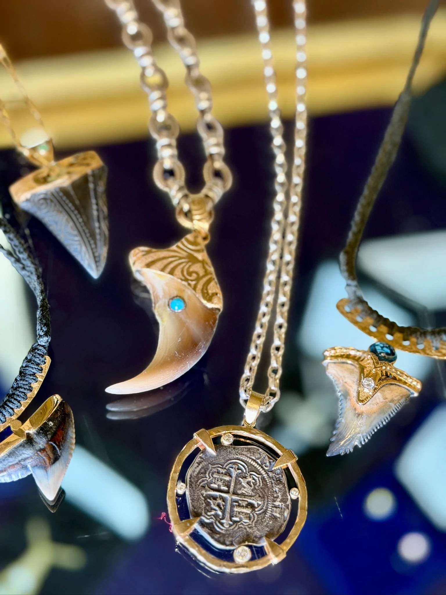 A display of gold jewelry necklaces and pendants, including a gold coin pendant with a cross and shield design, and shell-shaped charms with decorative patterns and blue gemstones.