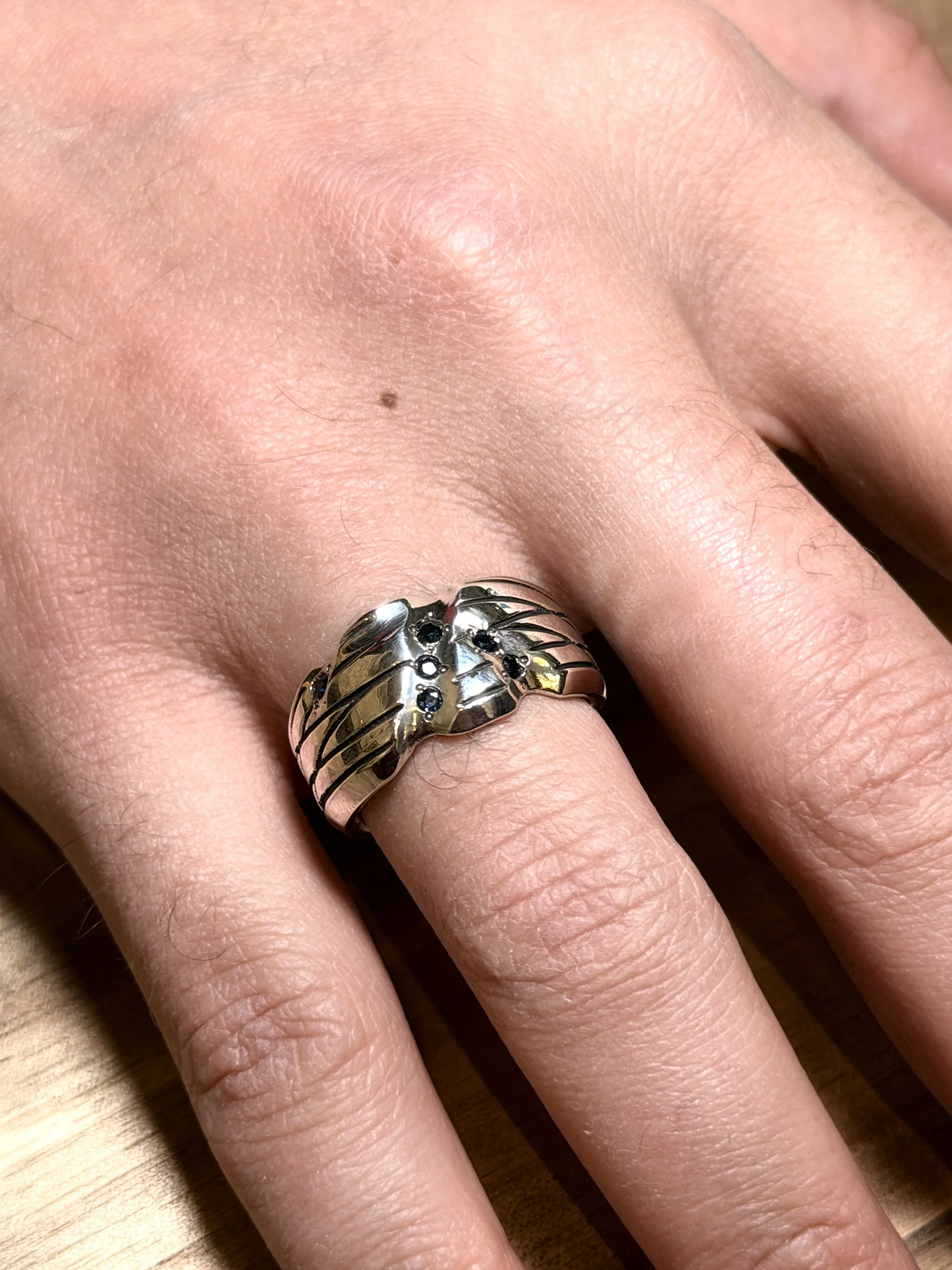 A hand with a silver ring shaped like a wing adorned with small dark gemstones, resting on a wooden surface.