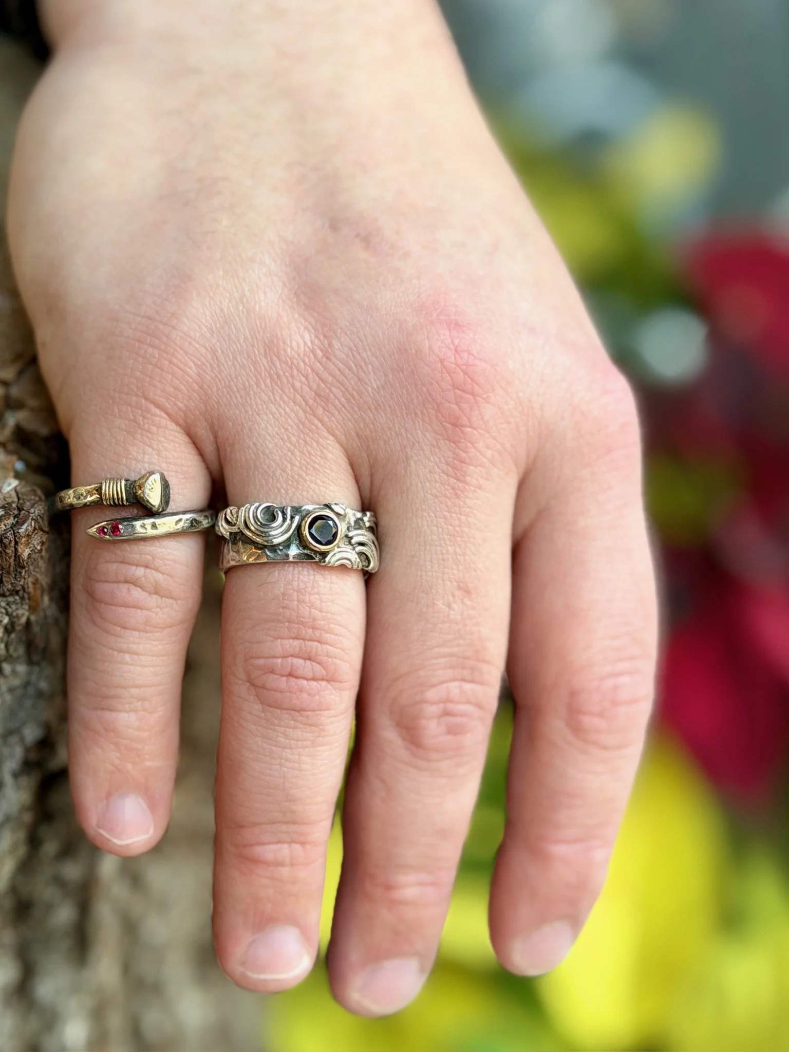 Close-up of a person's hand with three rings on fingers, positioned outdoors near a tree and blurred colorful background.