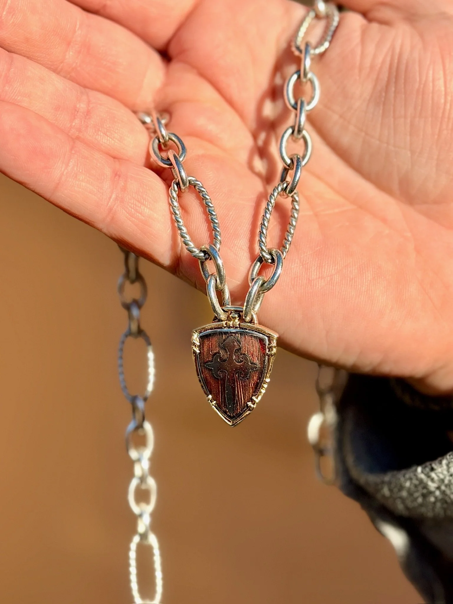A hand holds a silver necklace with a shield-shaped pendant featuring a cross and decorative elements.