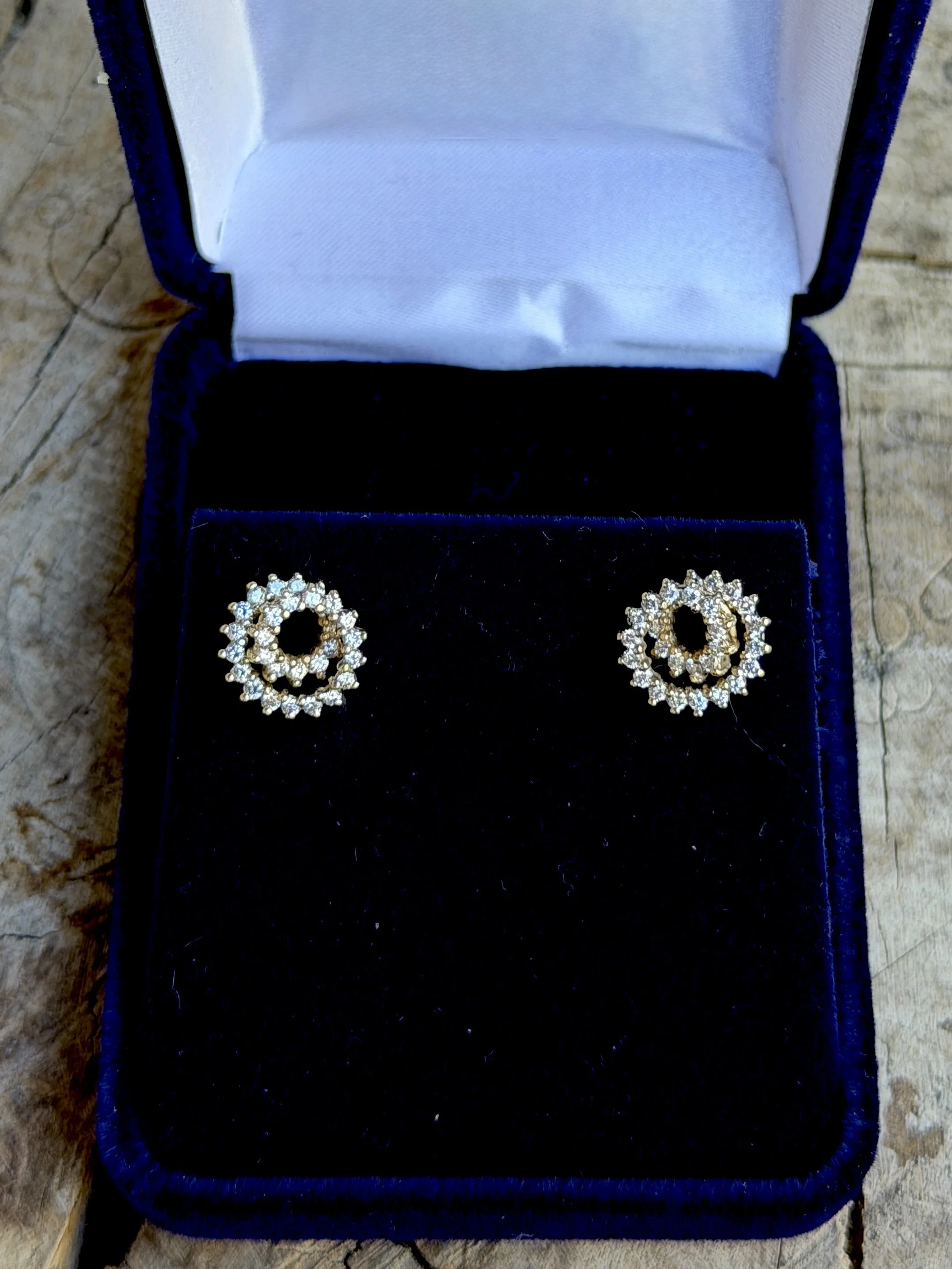 A pair of gold earrings with a circular design featuring multiple small diamonds, displayed inside an open dark blue jewelry box with a velvet interior.