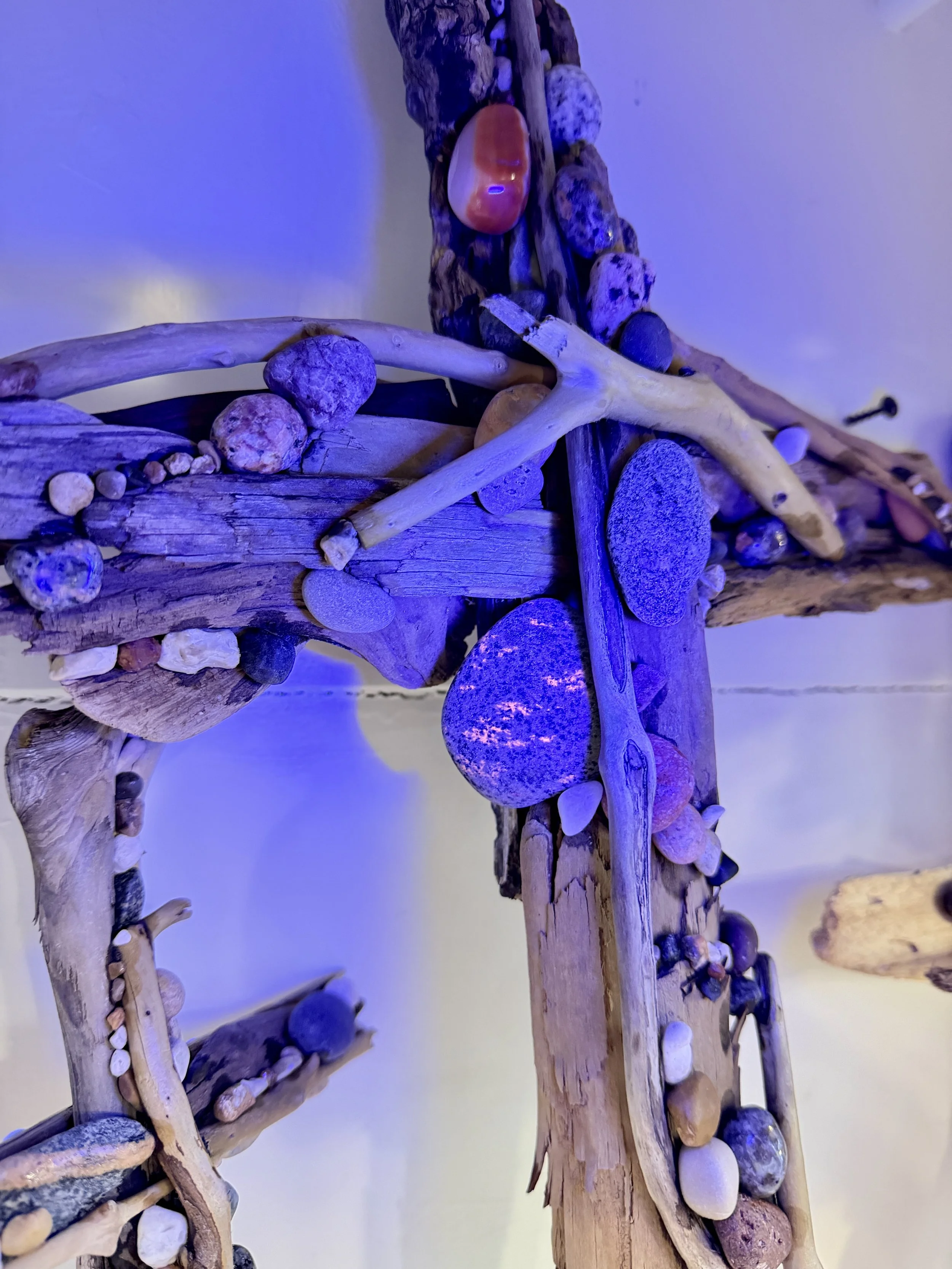 A wooden cross decorated with various small rocks and shells, illuminated with a blue light.