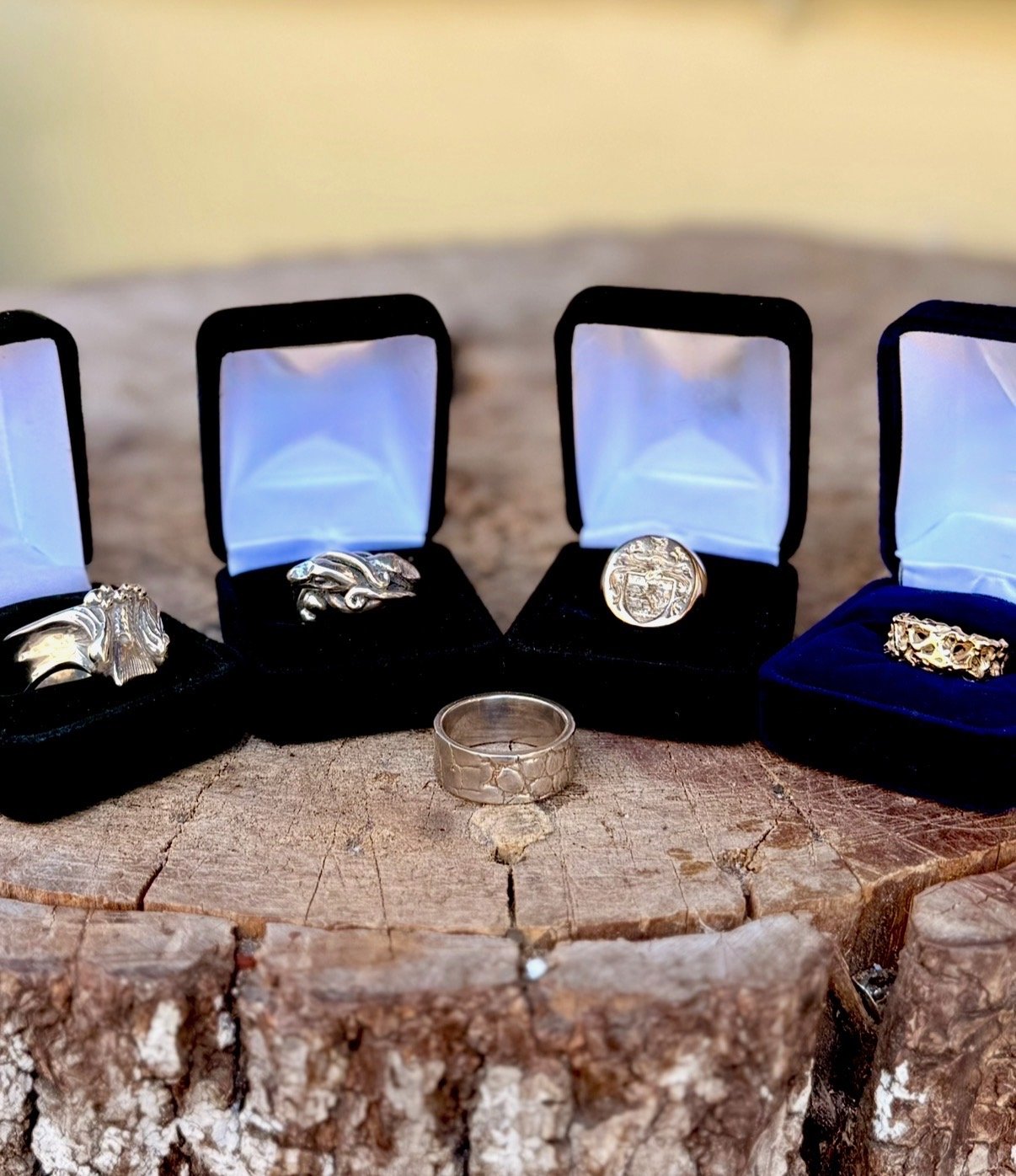 Four rings displayed on a wooden surface, with three rings in black velvet boxes and one ring outside the boxes. The rings are silver and gold with various designs.