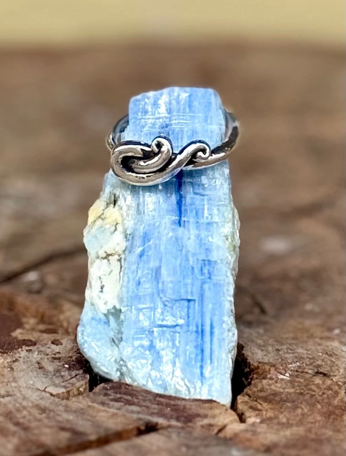 A silver ring with a design resembling a twisted infinity symbol, placed on a rough, light blue crystal or mineral on a wooden surface.