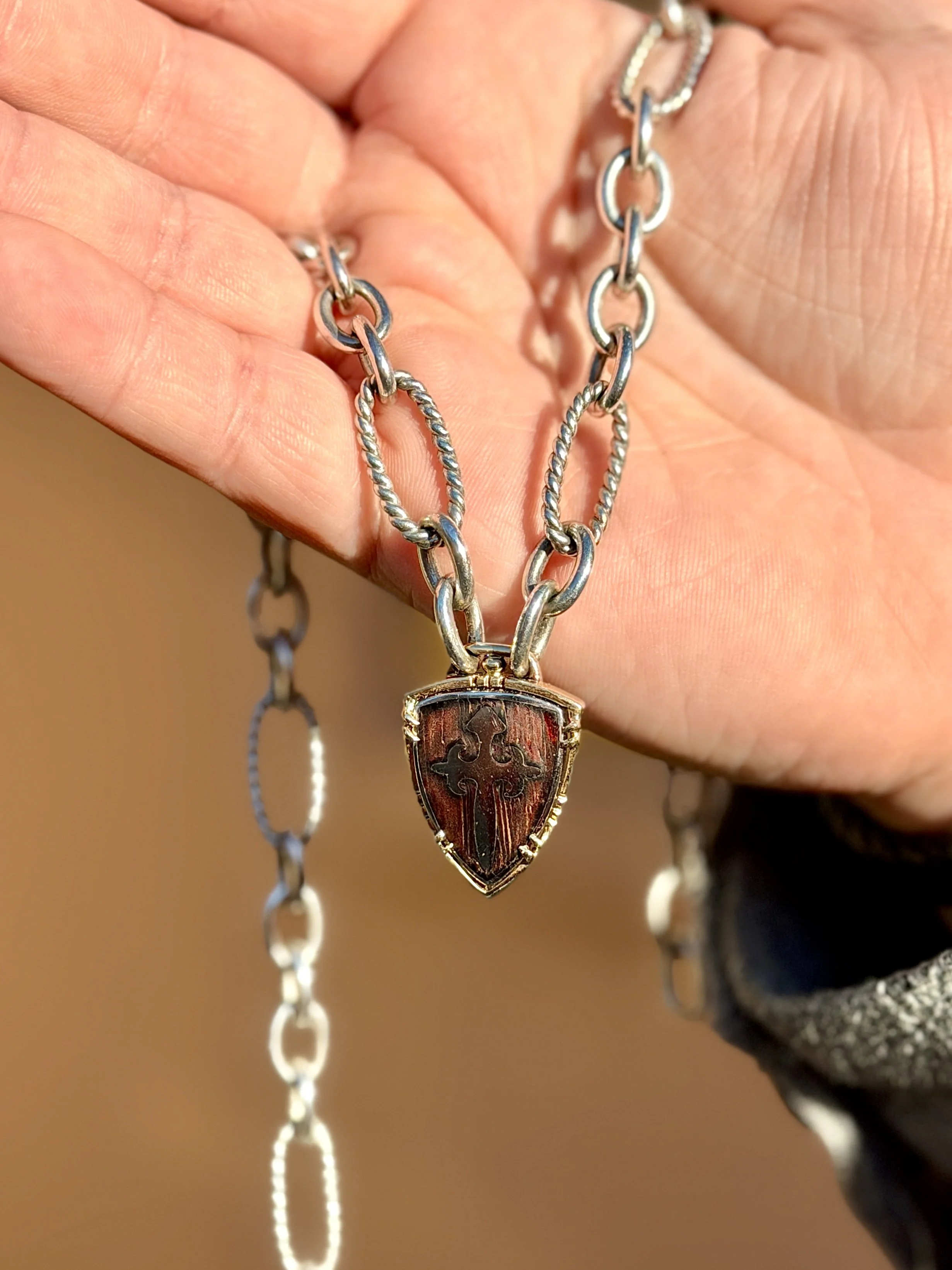 Close-up of a silver necklace with a shield-shaped pendant featuring a cross and a design resembling a fleur-de-lis.