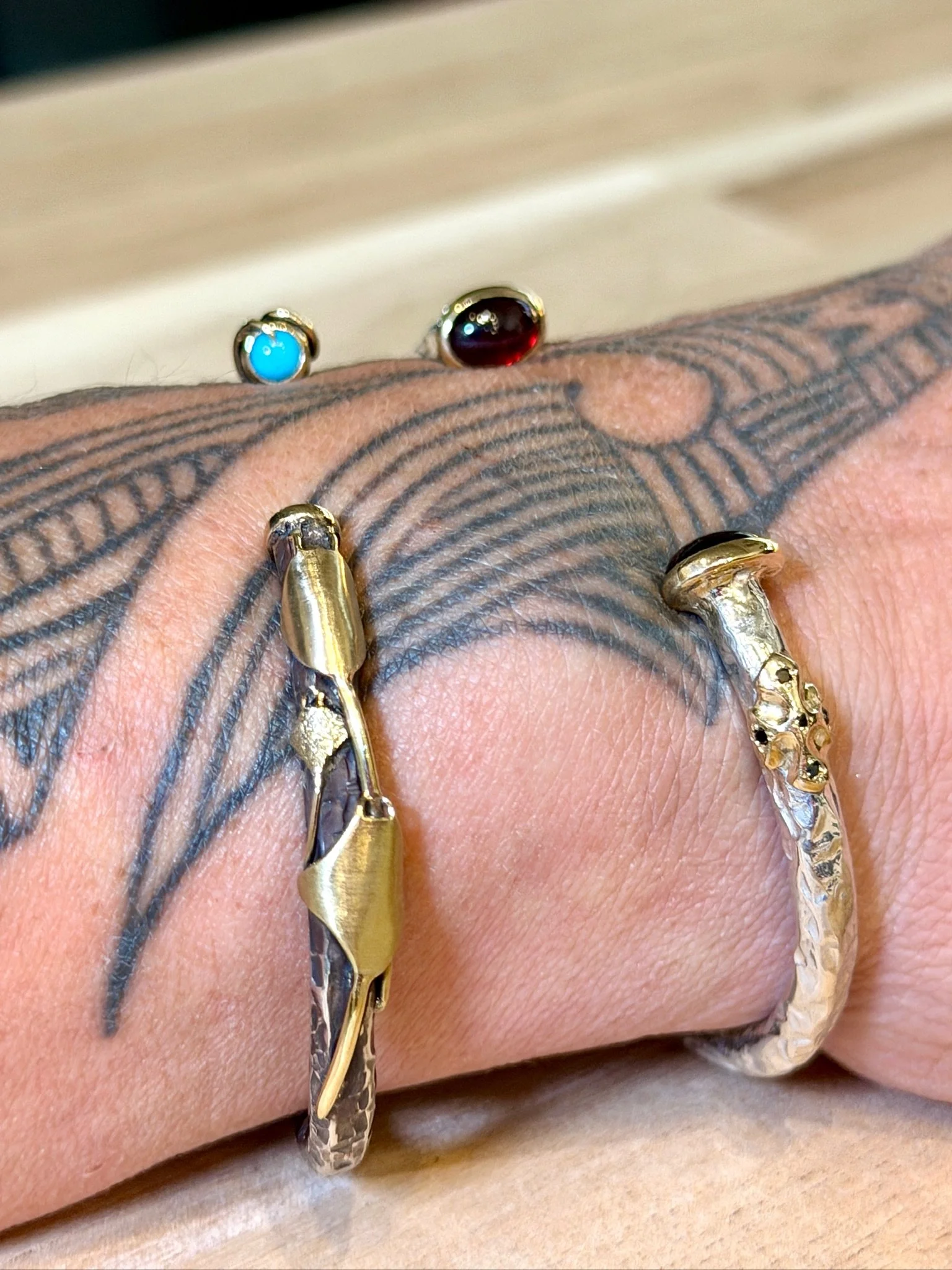 Close-up of four rings, two gold and two with colorful stones, worn on a hand with a black tattoo.