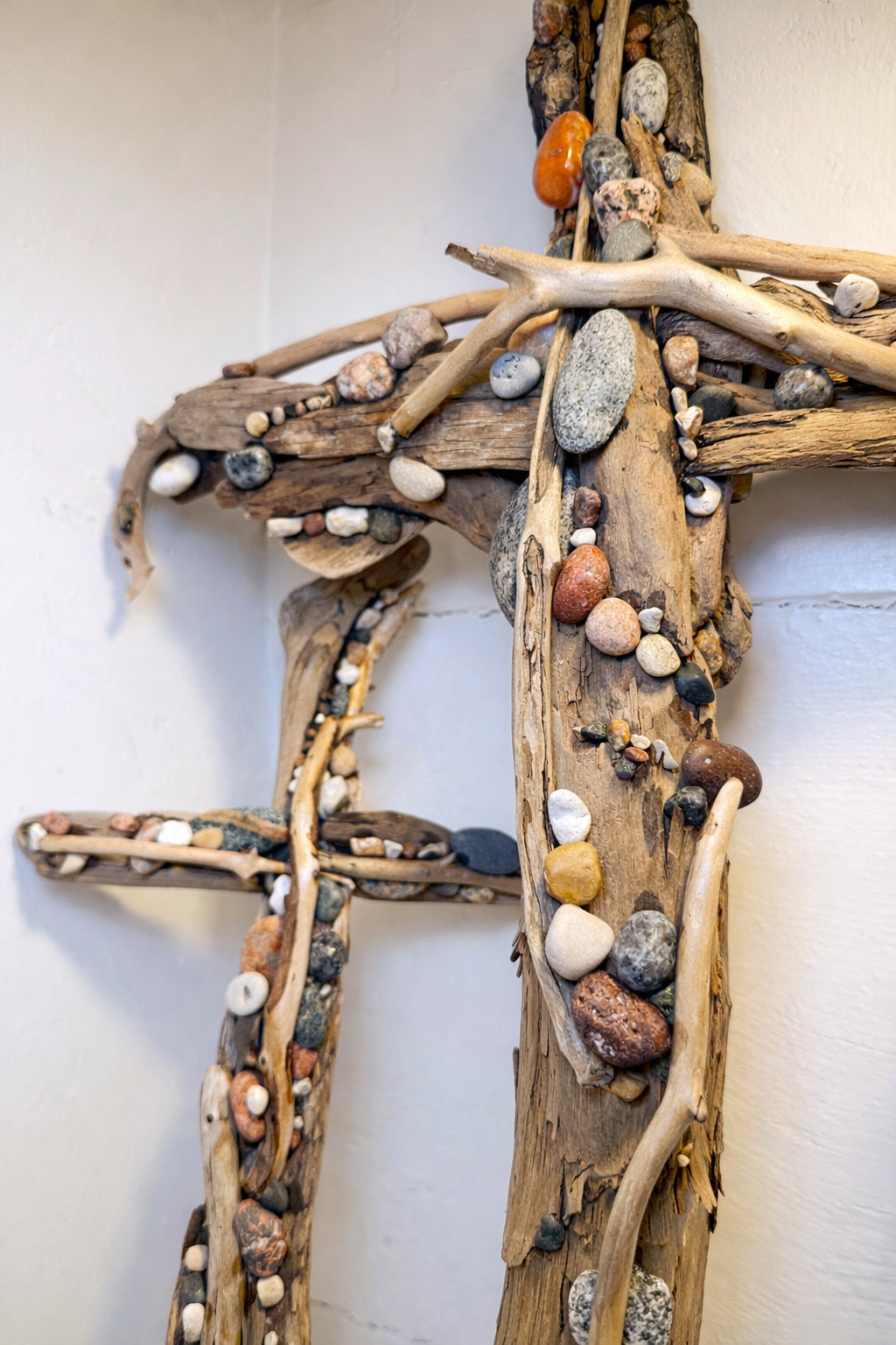 Decorative wall art made from driftwood and small colorful stones arranged in a cross shape on a white wall.