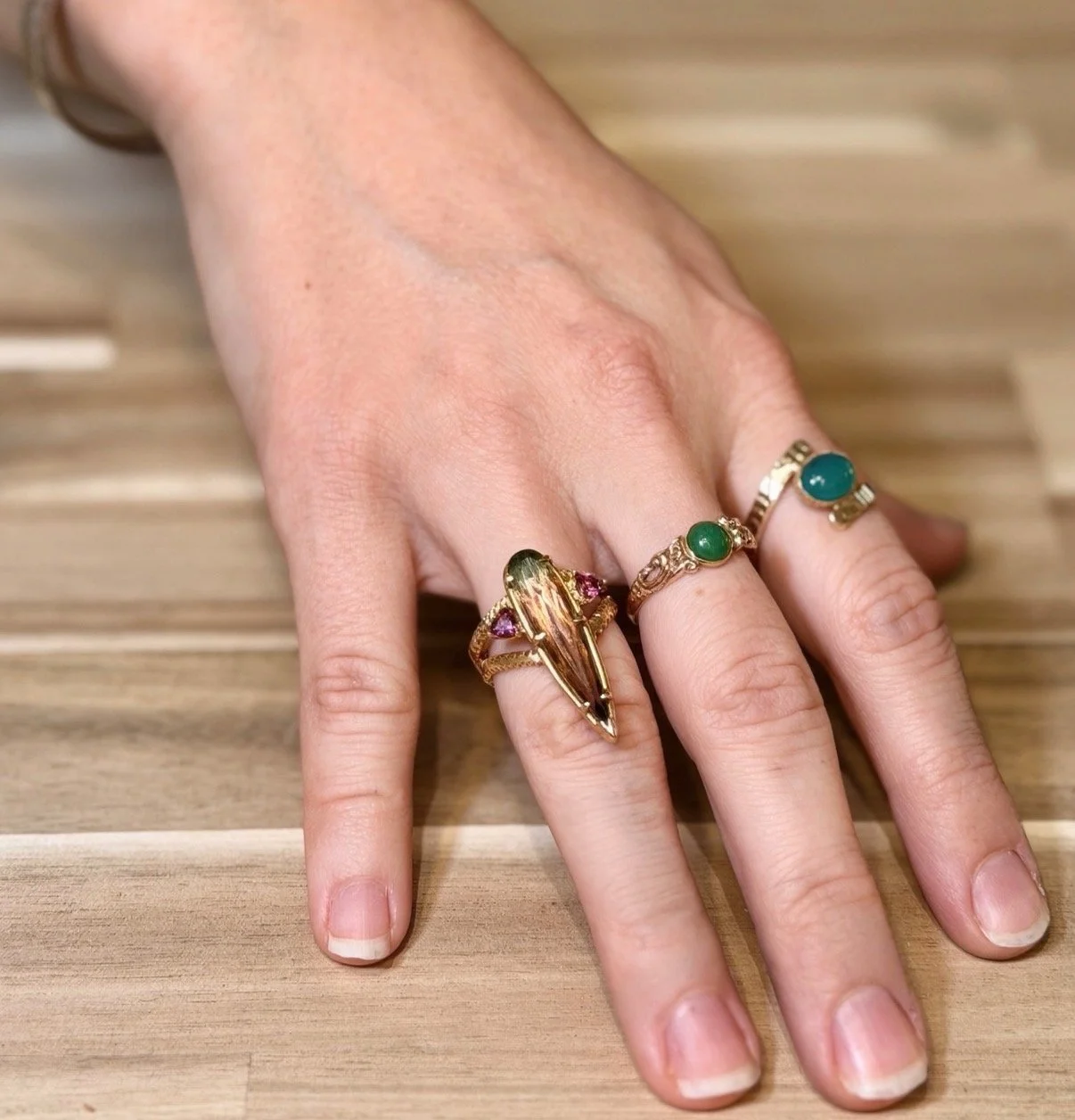 A hand displaying three rings with colorful stones on a wooden surface.