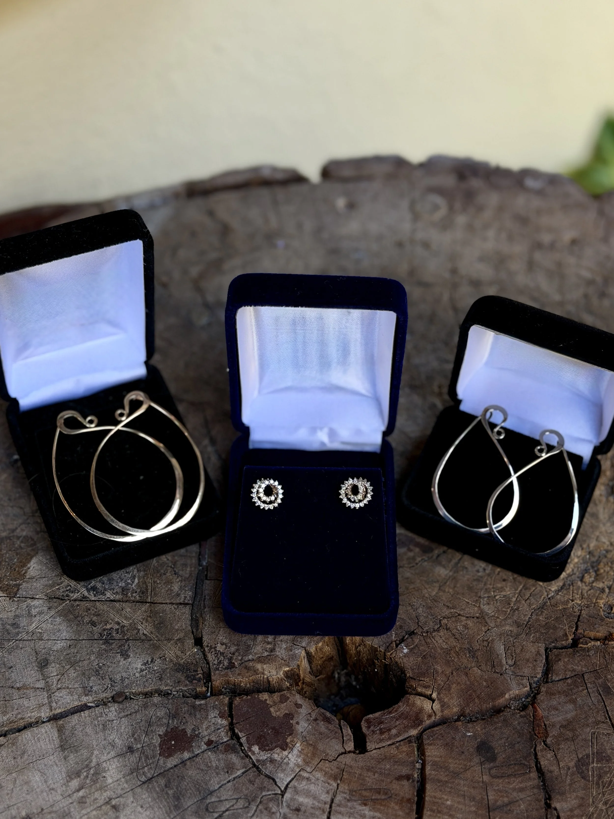 Three jewelry boxes with earrings inside, placed on a wooden surface. The left and right boxes contain hoop earrings, while the middle box contains a pair of circular earrings with gem accents.