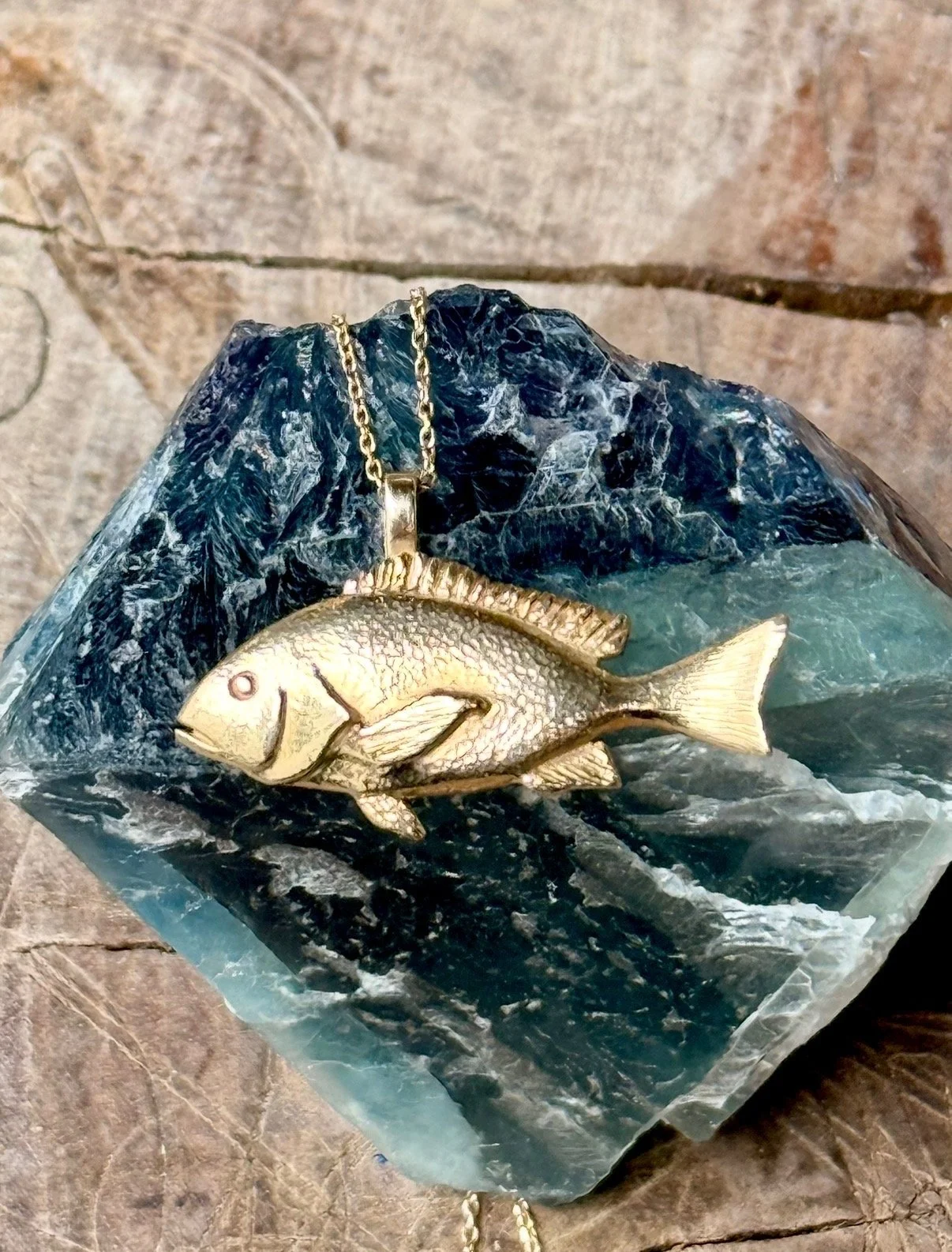A gold-colored fish pendant necklace resting on a large, dark, and light blue mineral or gemstone.