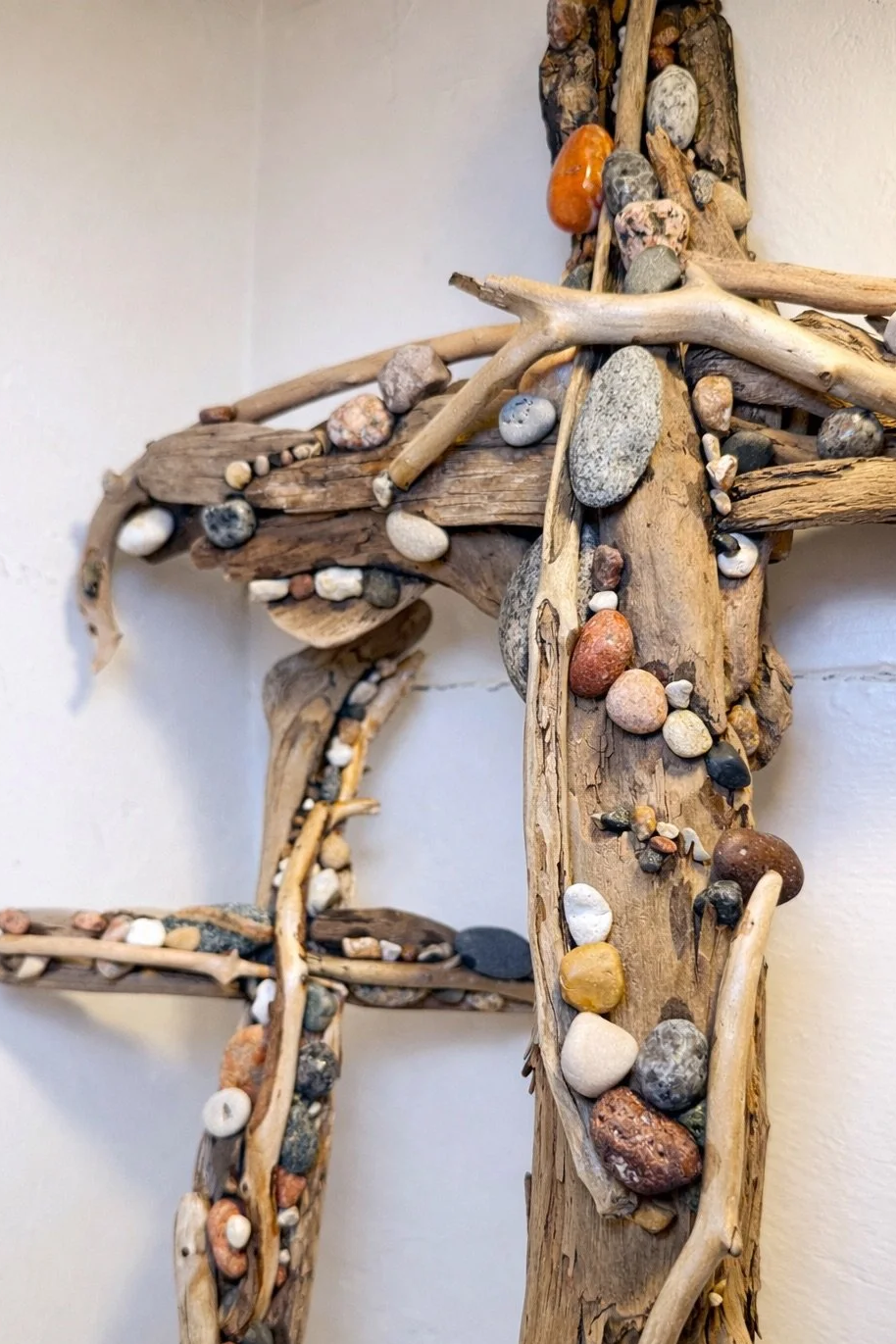 Wall-mounted decorative cross made of driftwood and various small stones.