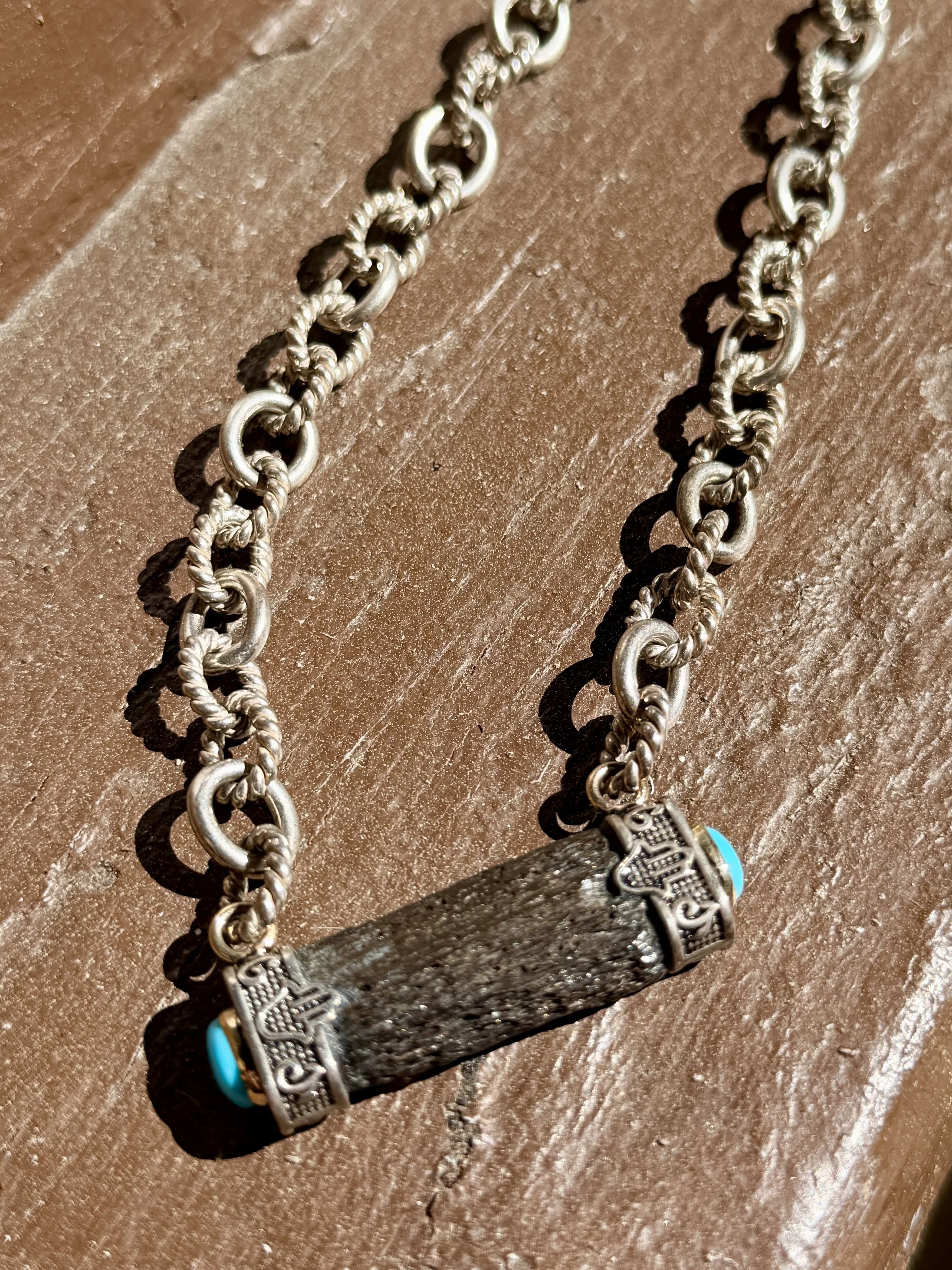 A necklace with a dark textured rectangular pendant and turquoise accents on both ends, resting on a wooden surface.