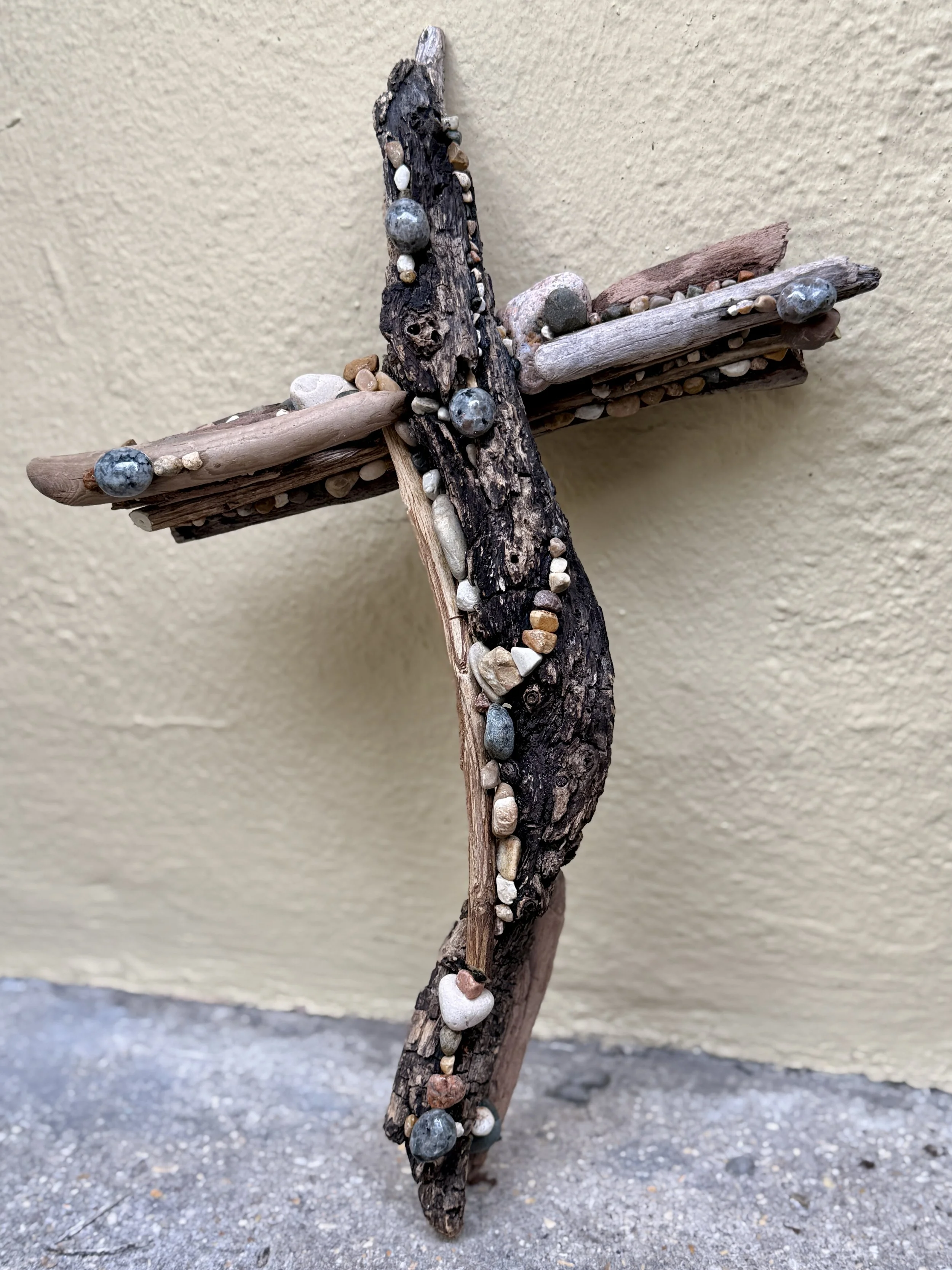 A cross made of driftwood and small pebbles, leaning against a beige wall.