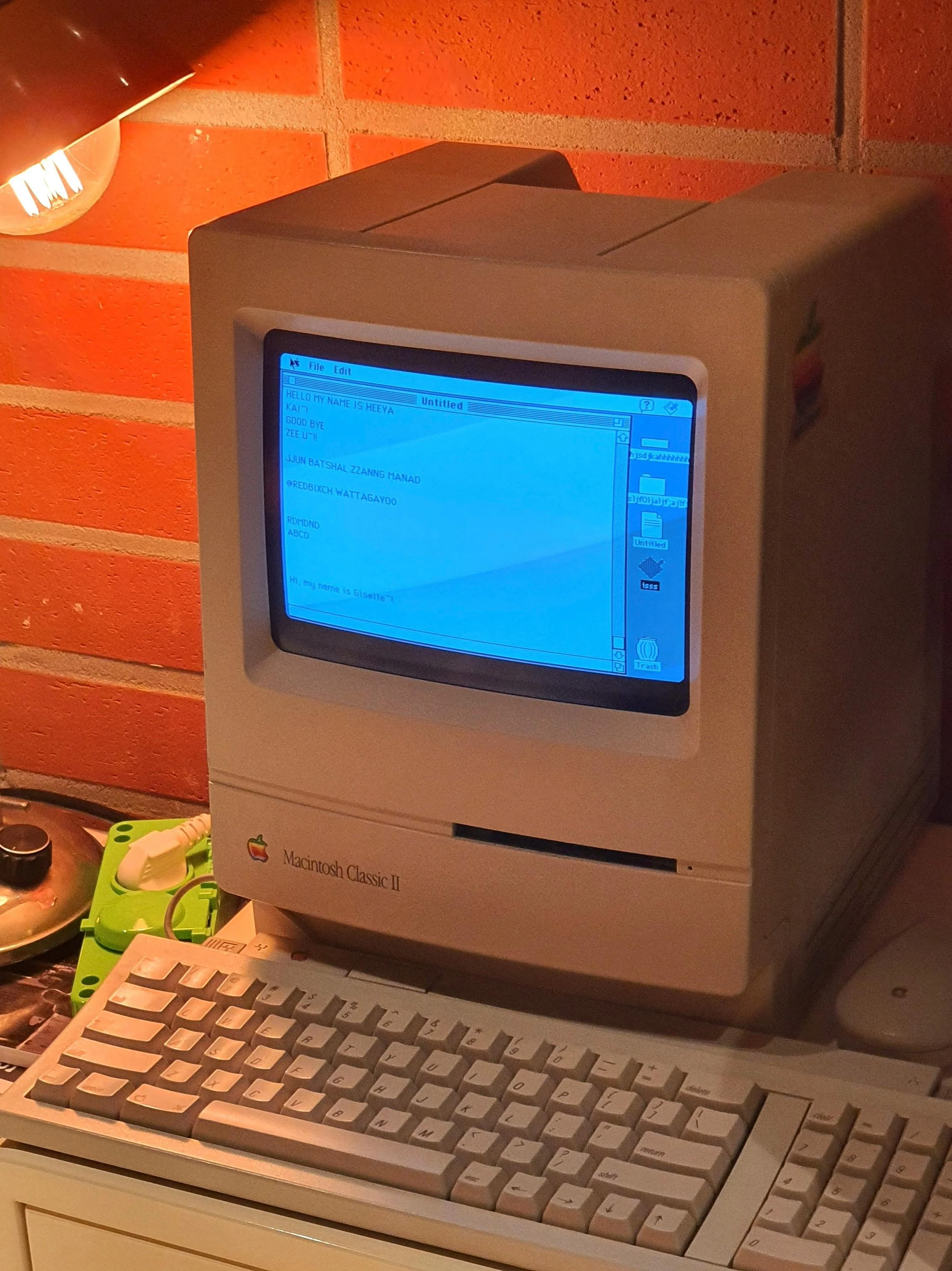 An old Apple Macintosh Classic II computer with a beige keyboard on a desk, against a brick wall background. The monitor displays a blue screen with text, and there are various small objects beside the computer, including a bowl with snack packets and a green container.