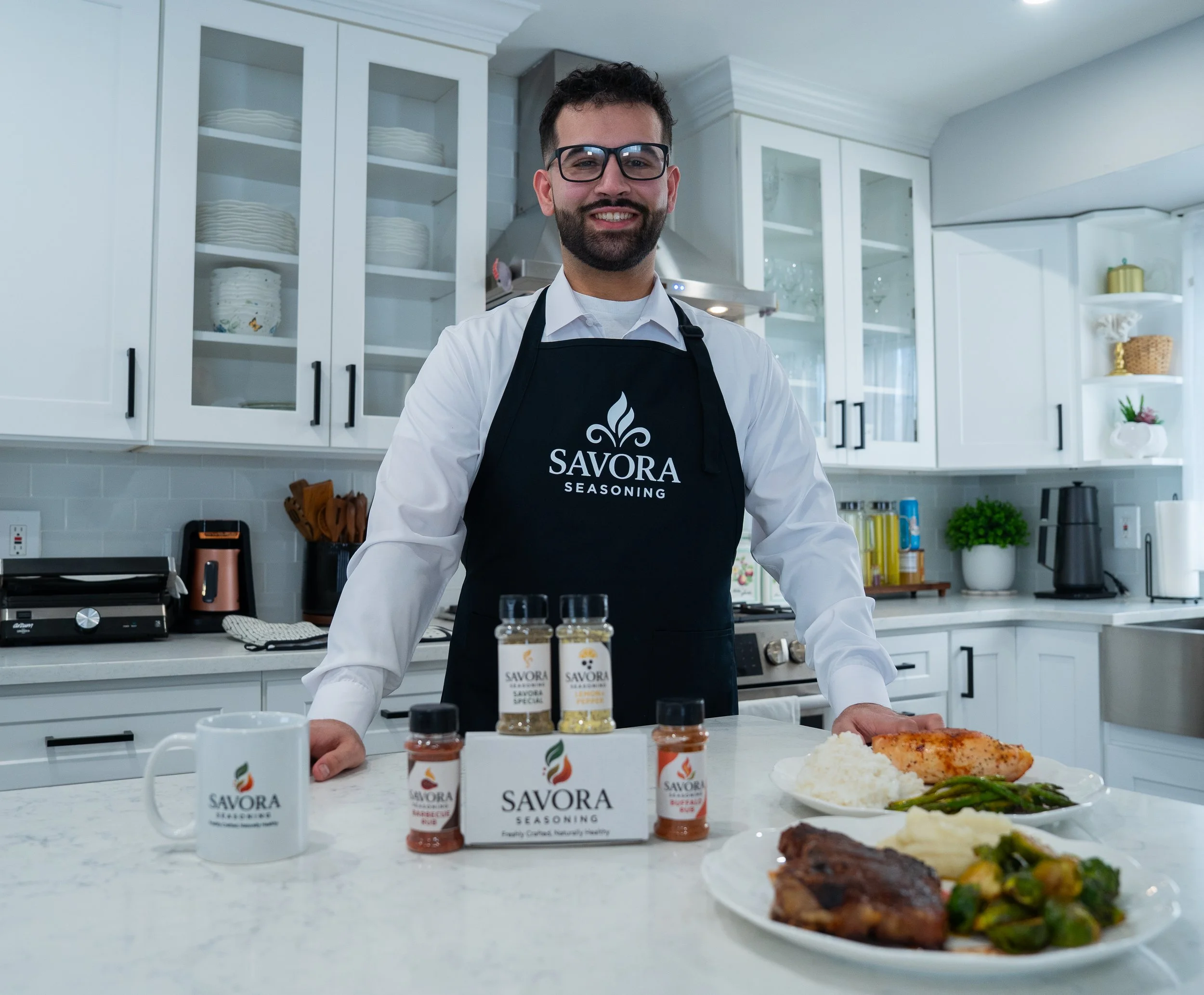A man smiling in a kitchen, wearing glasses and a black apron with Savora Seasoning logo, standing behind a counter with spices and dishes of food.