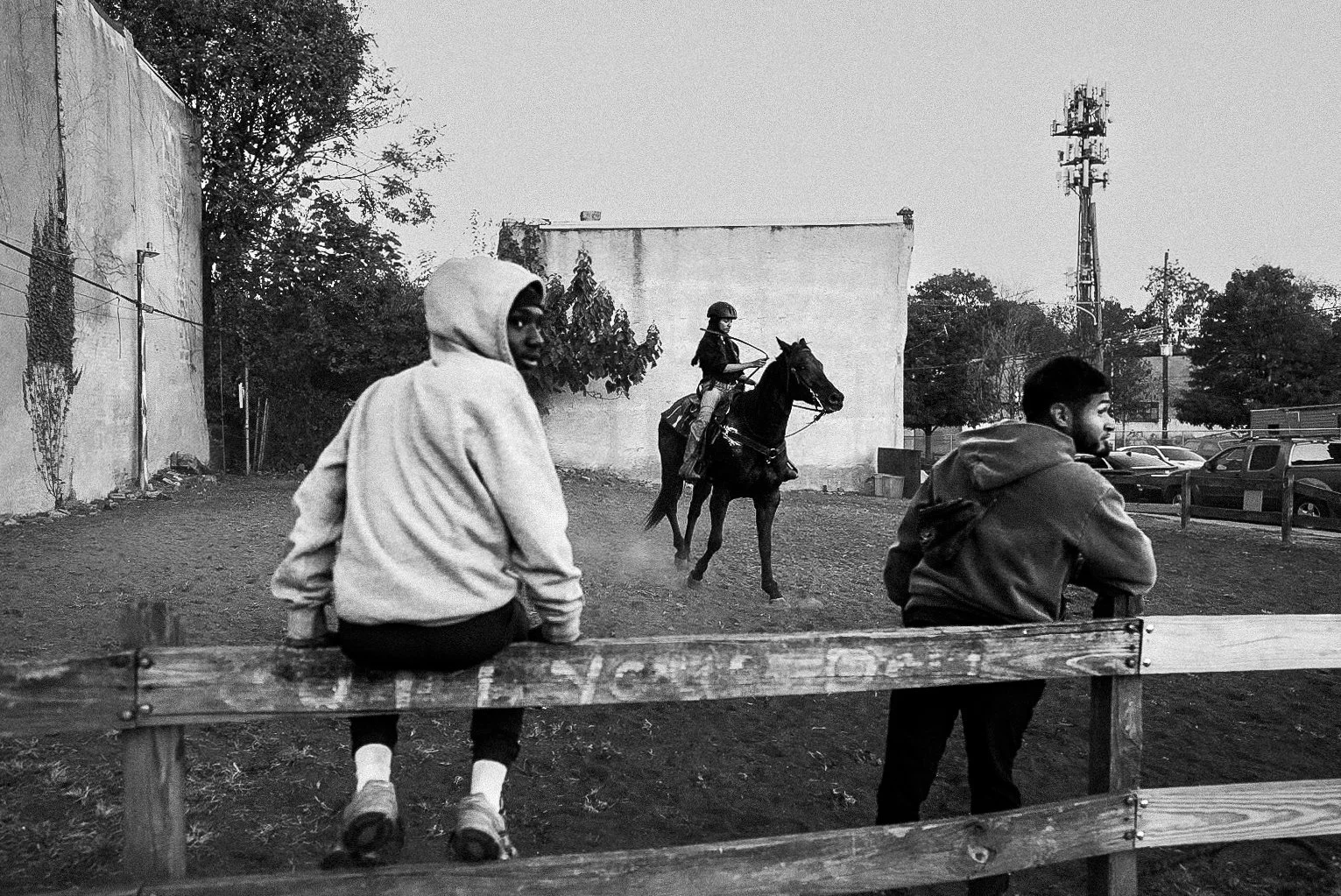 Two young people sitting on a wooden fence and a person riding a horse in an outdoor area, with trees, a wall, cars, and a utility pole in the background, captured in black and white.