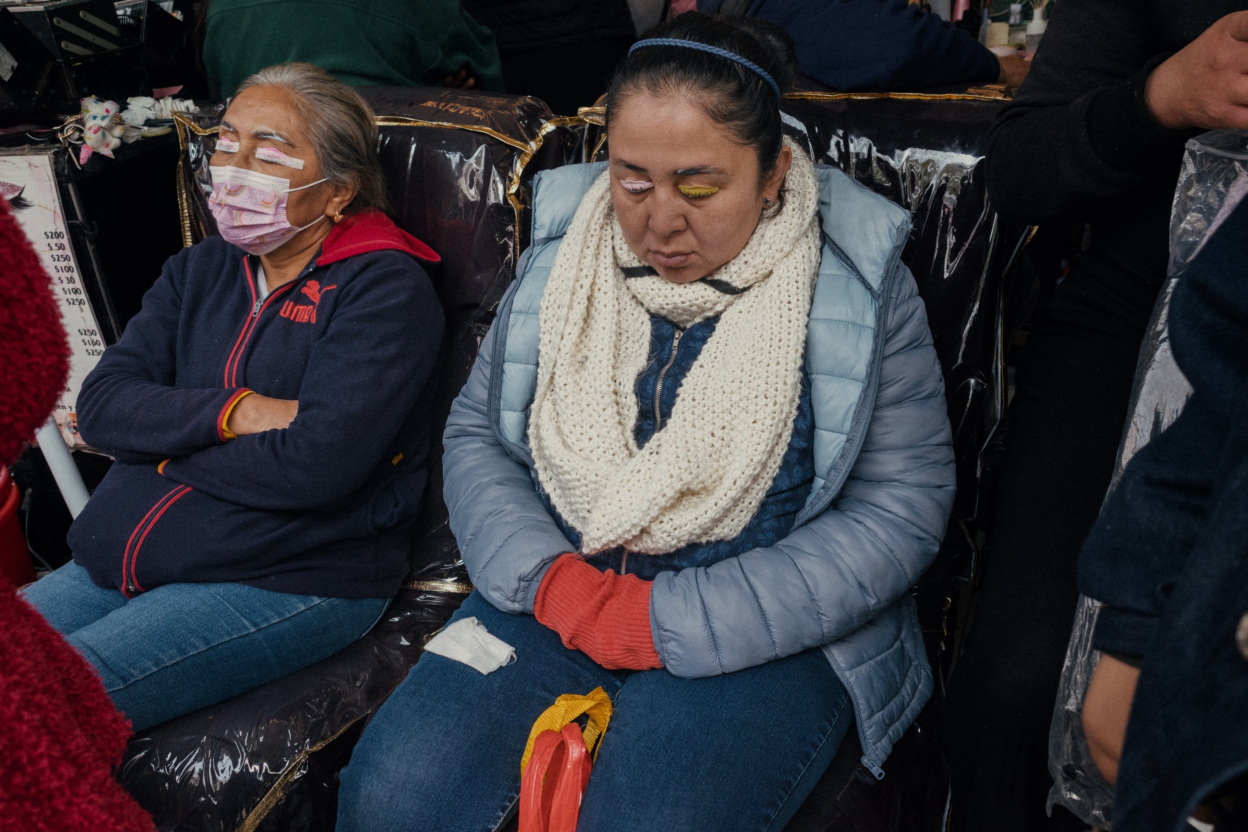 Two women sitting with eyes closed, wearing warm clothing, on a crowded bus or train, with some people standing nearby.