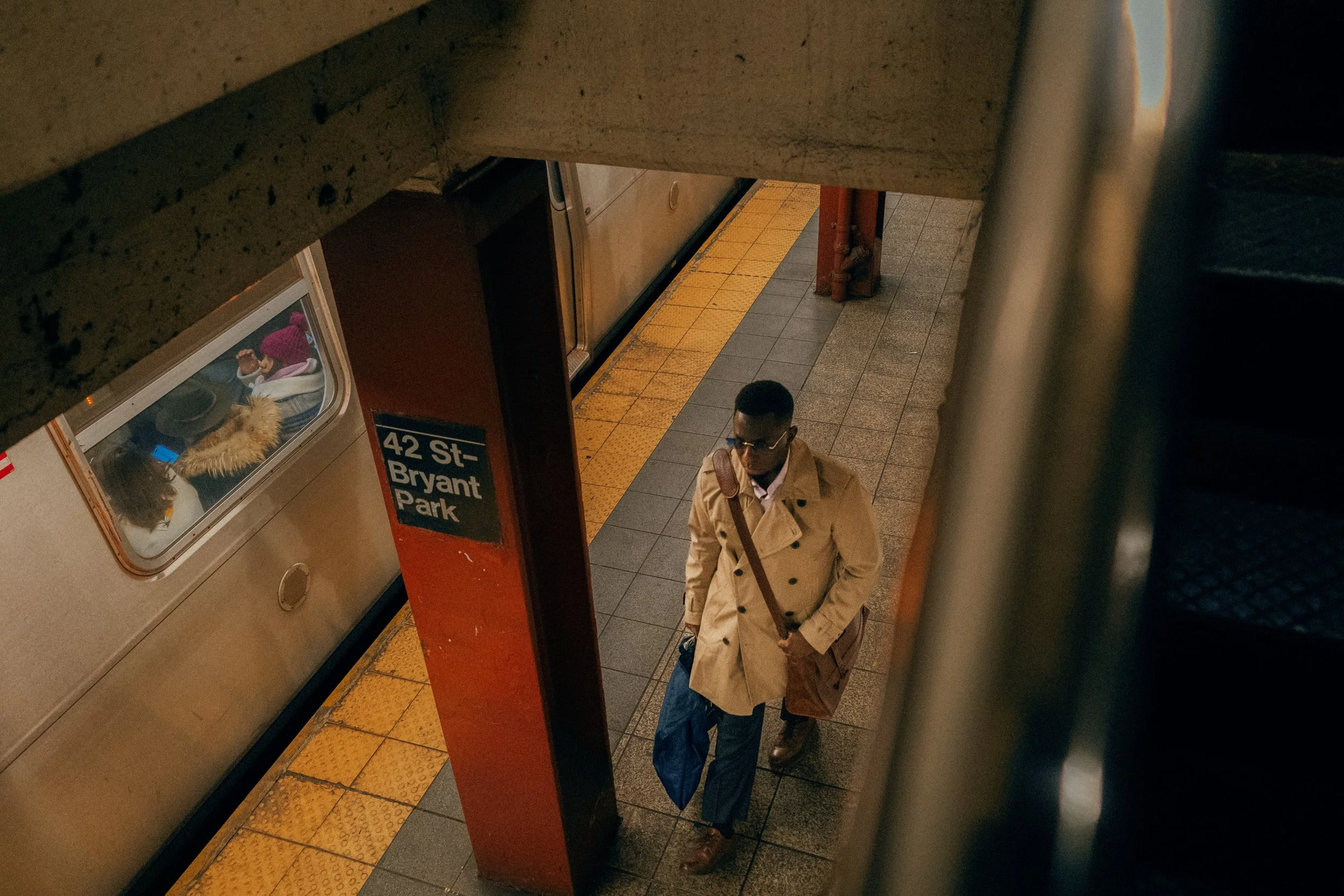 A man in a beige trench coat carrying a shoulder bag and a blue umbrella walking on the subway platform at 42 St-Bryant Park station, seen from above.