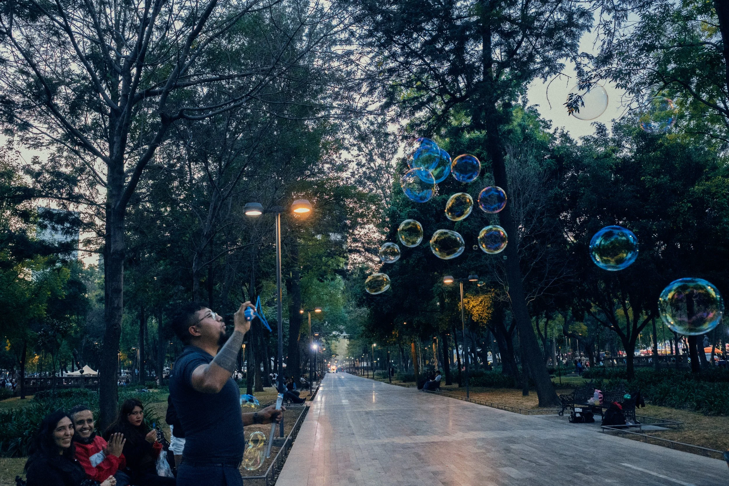 A man blowing soap bubbles in a park during twilight while three women sit on a bench and watch him, with trees and streetlights in the background.