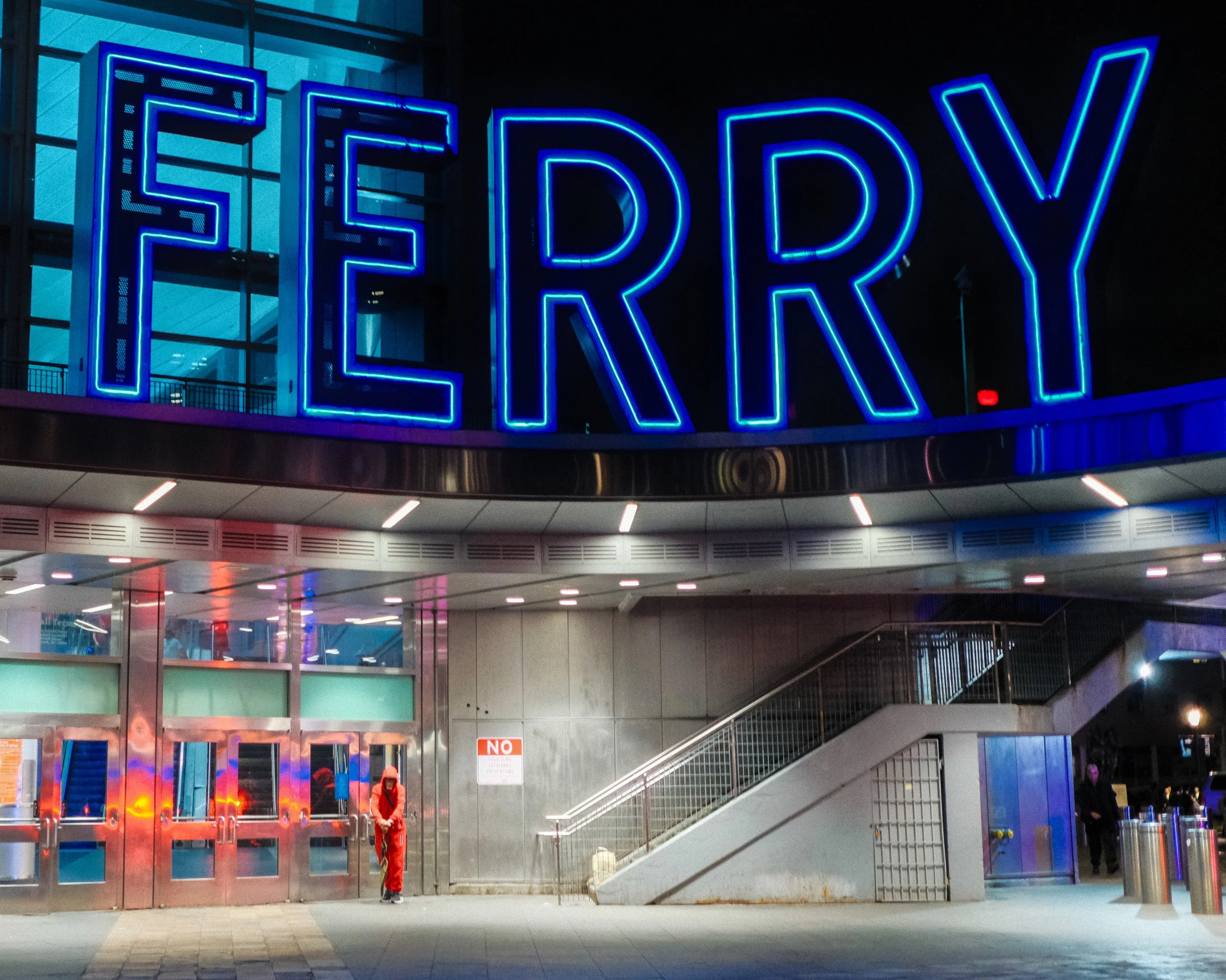 Nighttime scene of a building with a large blue neon sign that reads 'FERRY.' A person in orange clothing stands near the entrance, and there are a few other people on the right side of the image. The building has glass doors and stairs leading up to