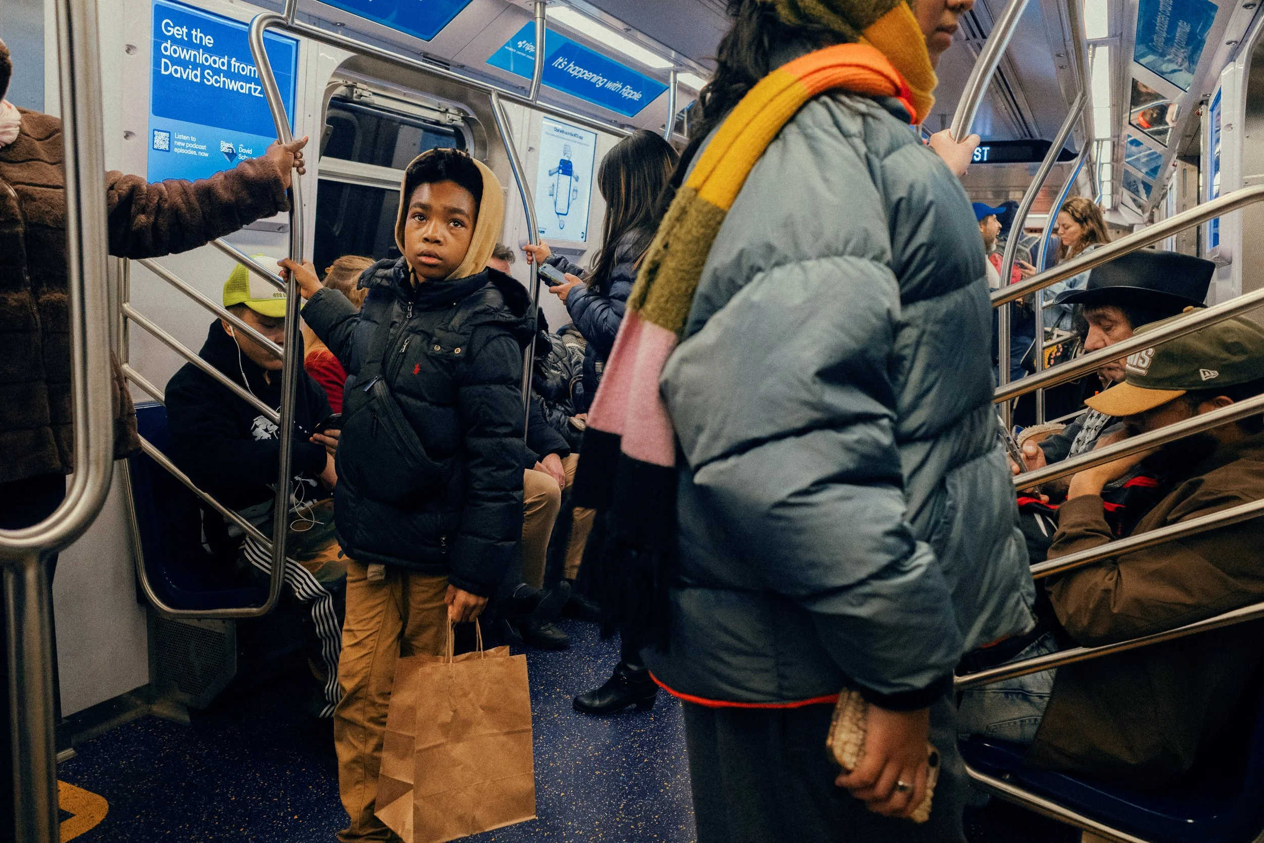 A crowded subway train with passengers standing and sitting, including a young boy holding a paper bag looking at the camera.