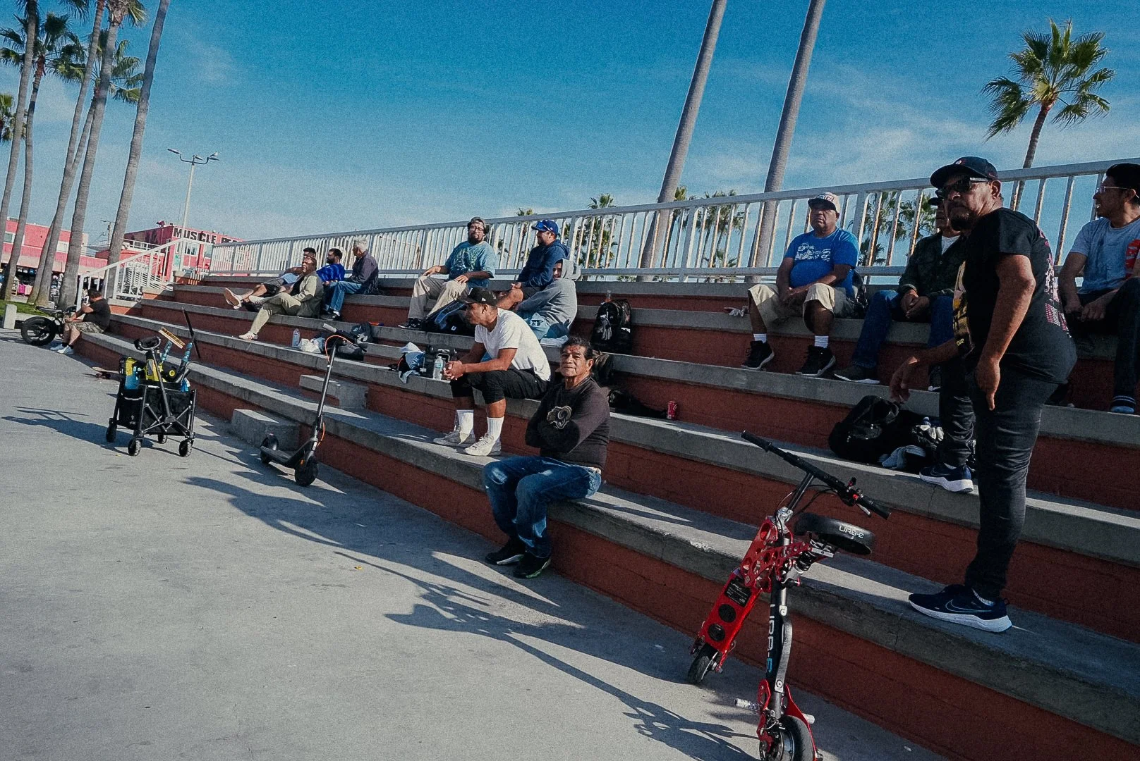People sitting on outdoor bleachers near palm trees on a sunny day, with electric scooters parked on the ground.
