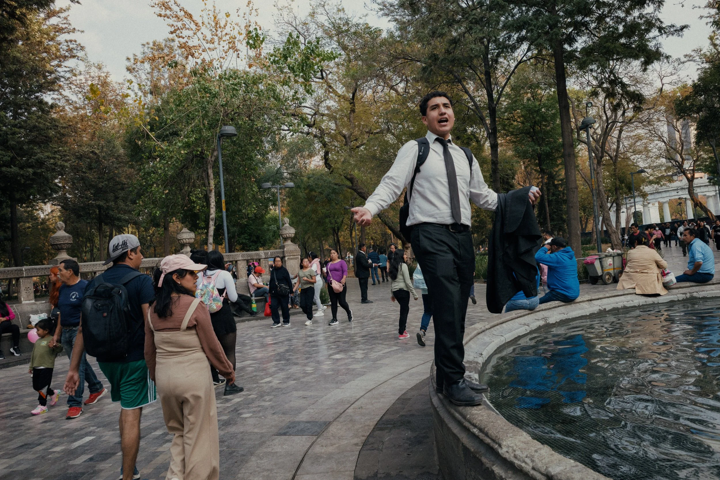 A man in a white shirt and black tie standing on the edge of a fountain, appearing to shout or sing, with a crowd of people in a park behind him.