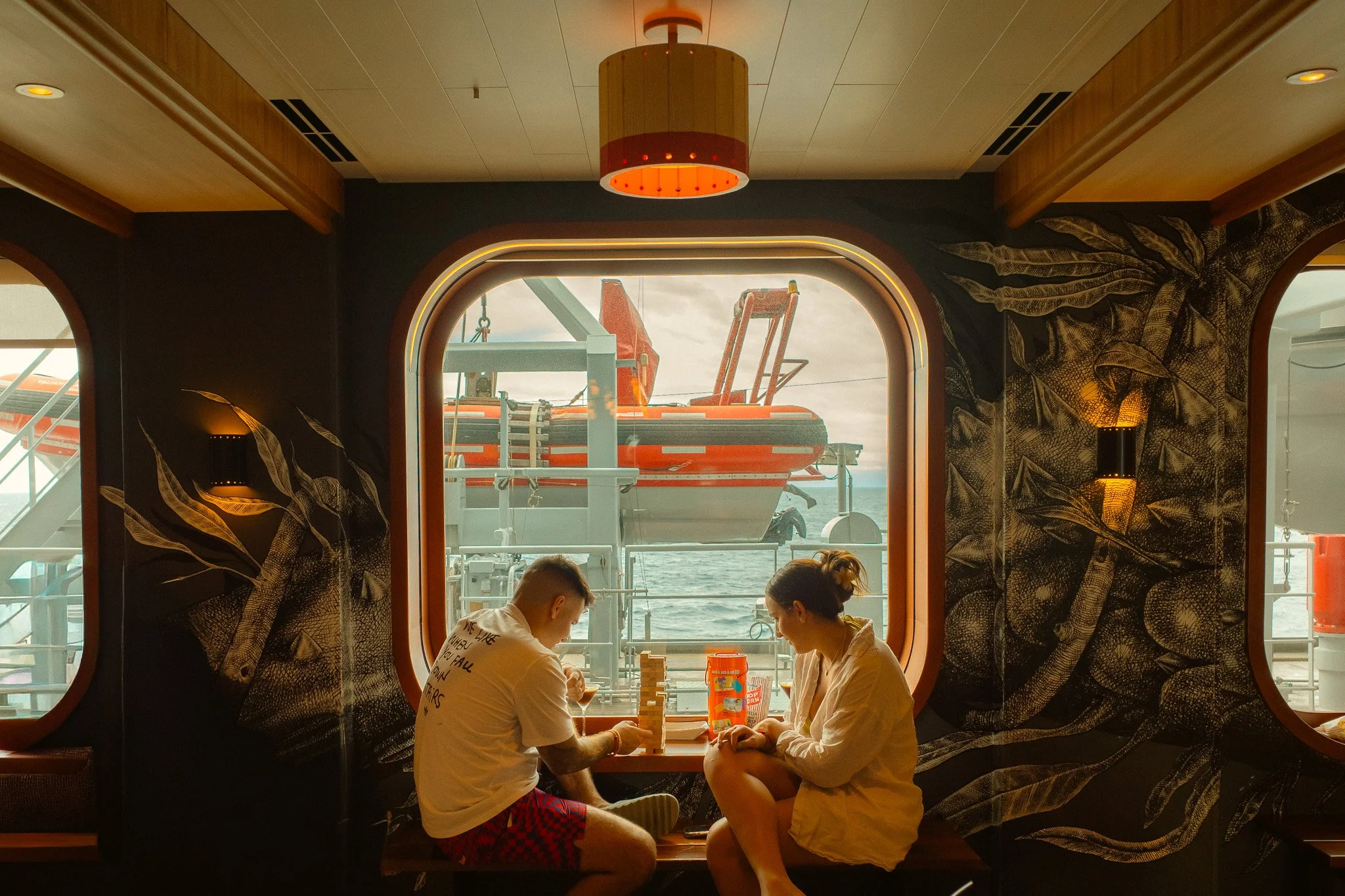 Two people sitting at a table on a cruise ship, playing Jenga, with an ocean view and a lifeboat outside the window.