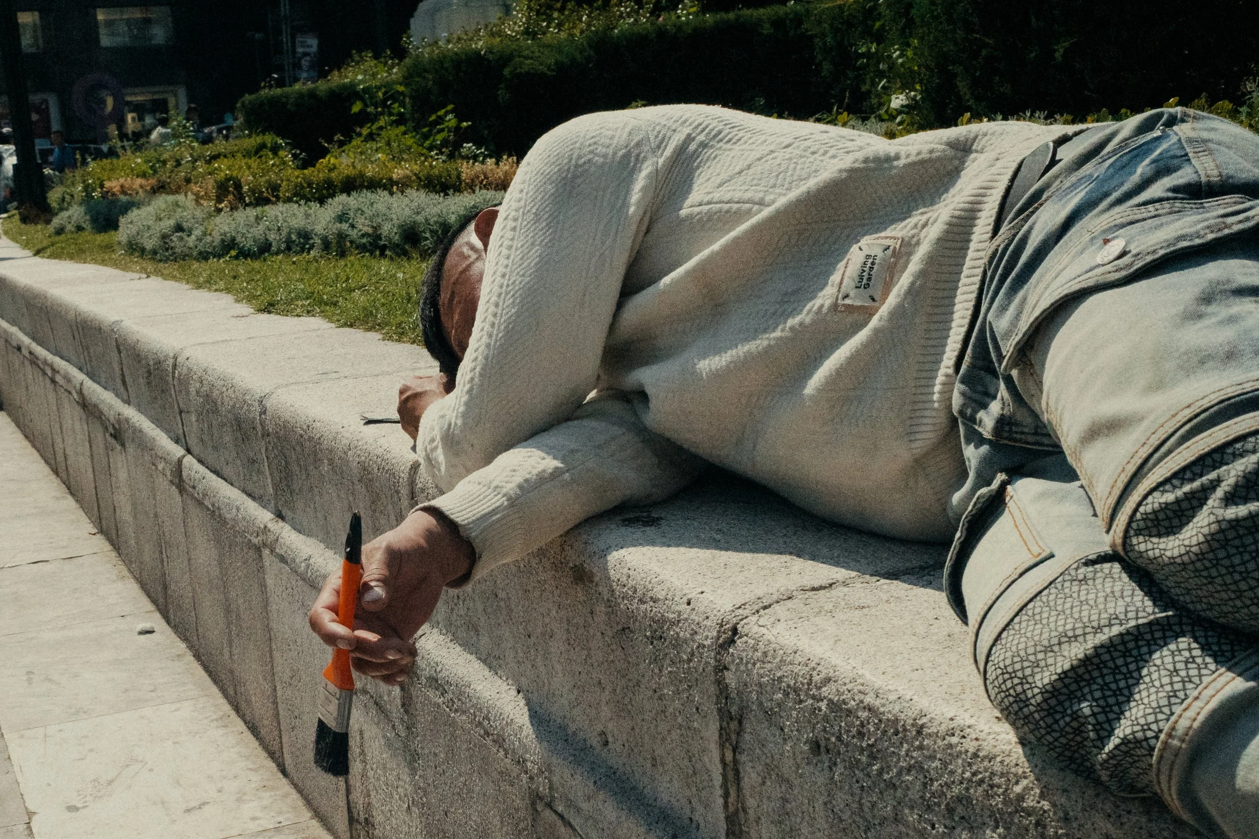 A person lying on a concrete sidewalk with their head and arm resting on a low wall, holding a paintbrush in their hand, wearing a white sweater and denim shorts, with greenery and a street in the background.
