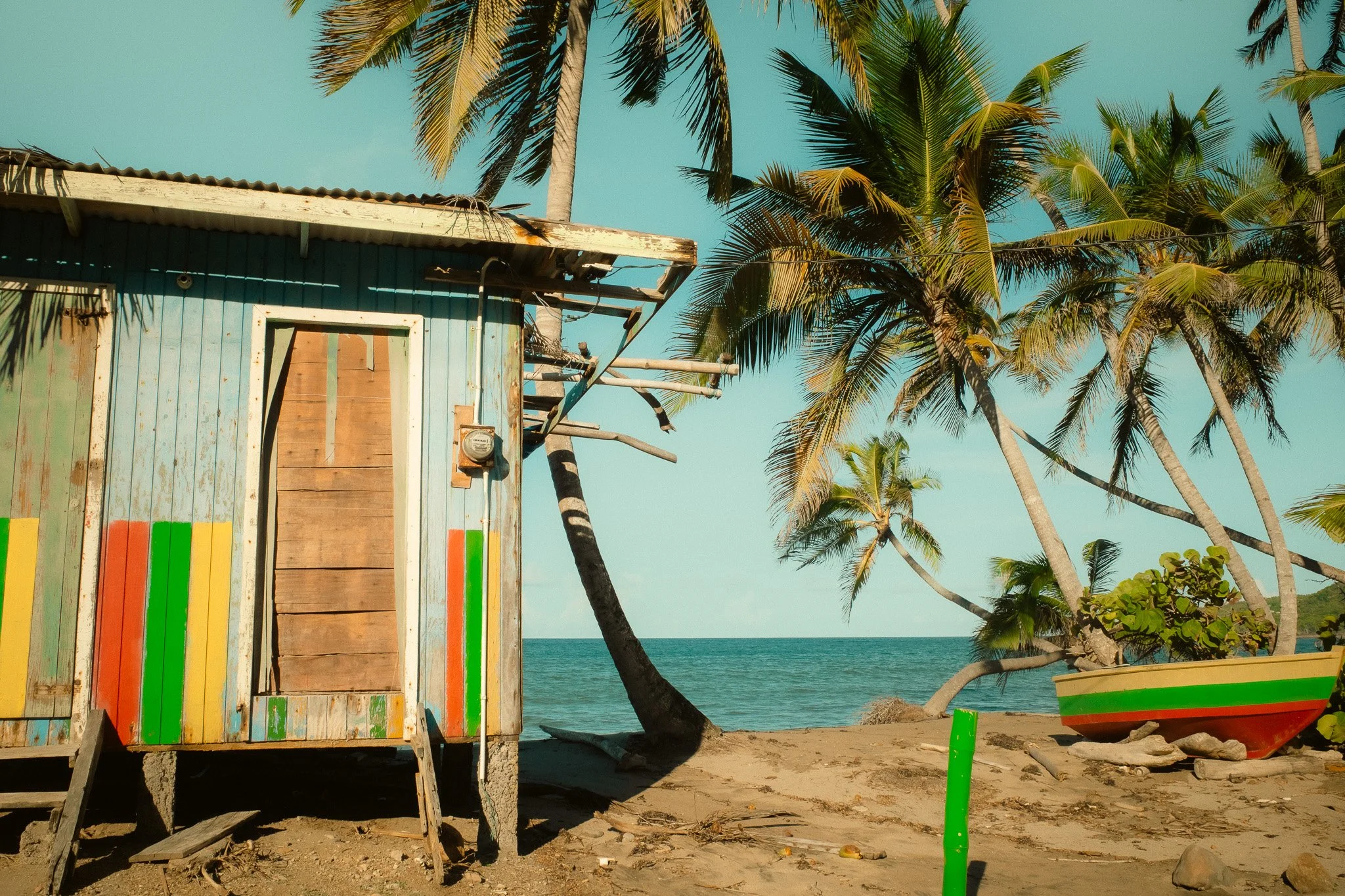 A weathered, multicolored beach hut on the sand with a small set of stairs leading up to its door, surrounded by tall palm trees and a boat painted with red, yellow, and green, seen on the right side, near the shoreline with the ocean in the backgrou