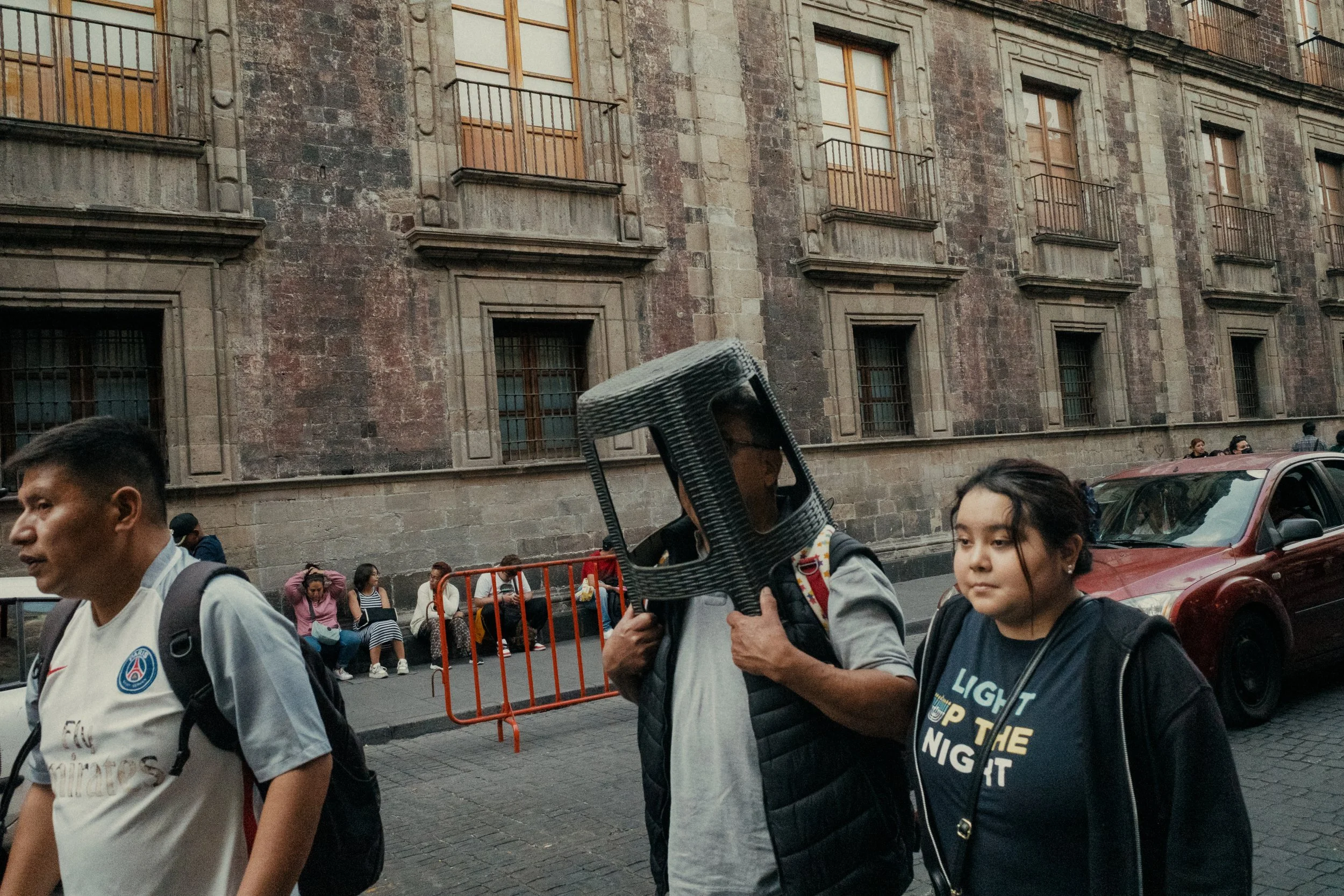 People walking on a city street with a historic stone building in the background, and a man carrying a carbon fiber mask.
