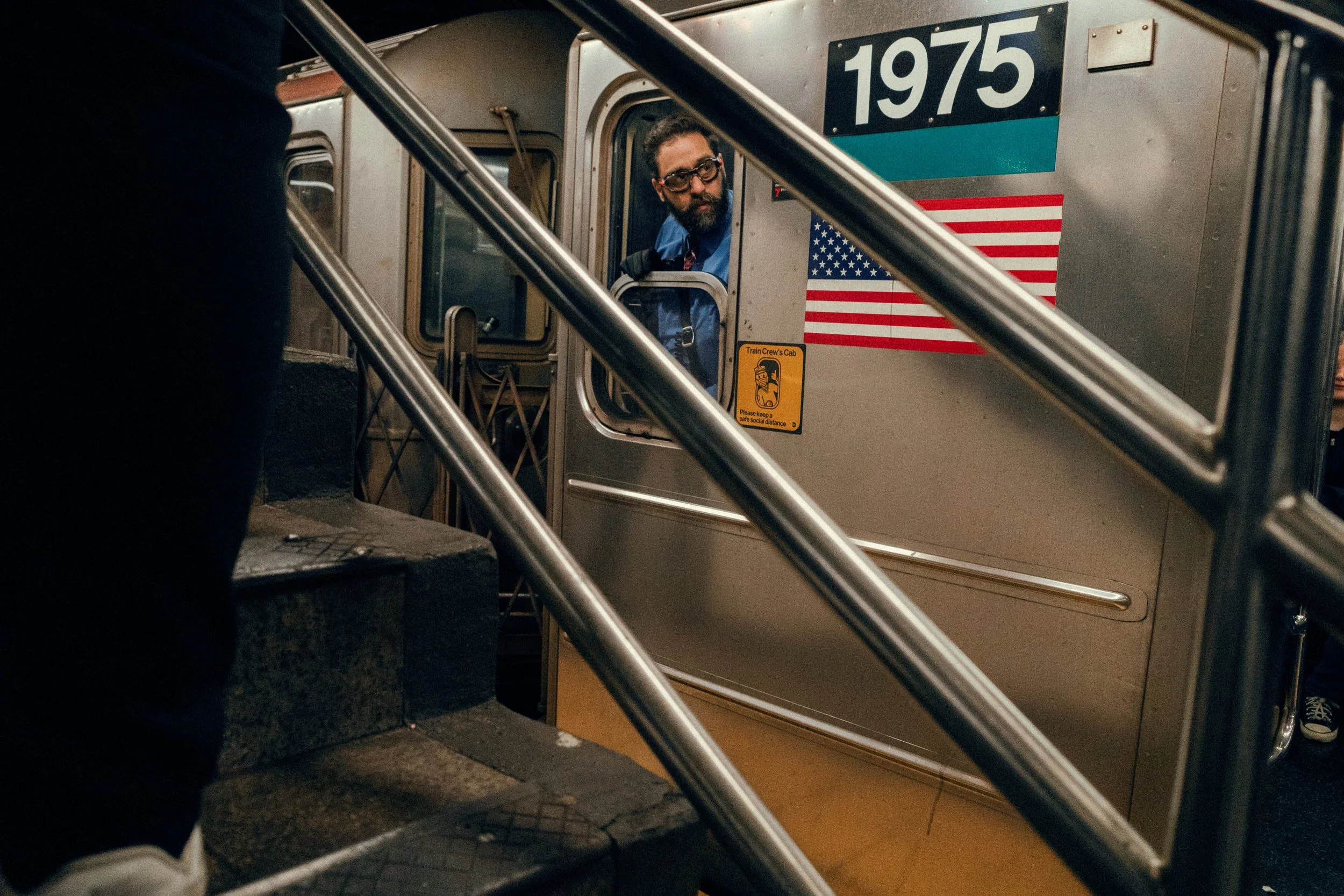 A man with a beard and glasses looks out from the entrance of a 1975 subway train, which has an American flag and a yellow safety sign on its exterior. The photo is taken from the stairs leading up to the train.