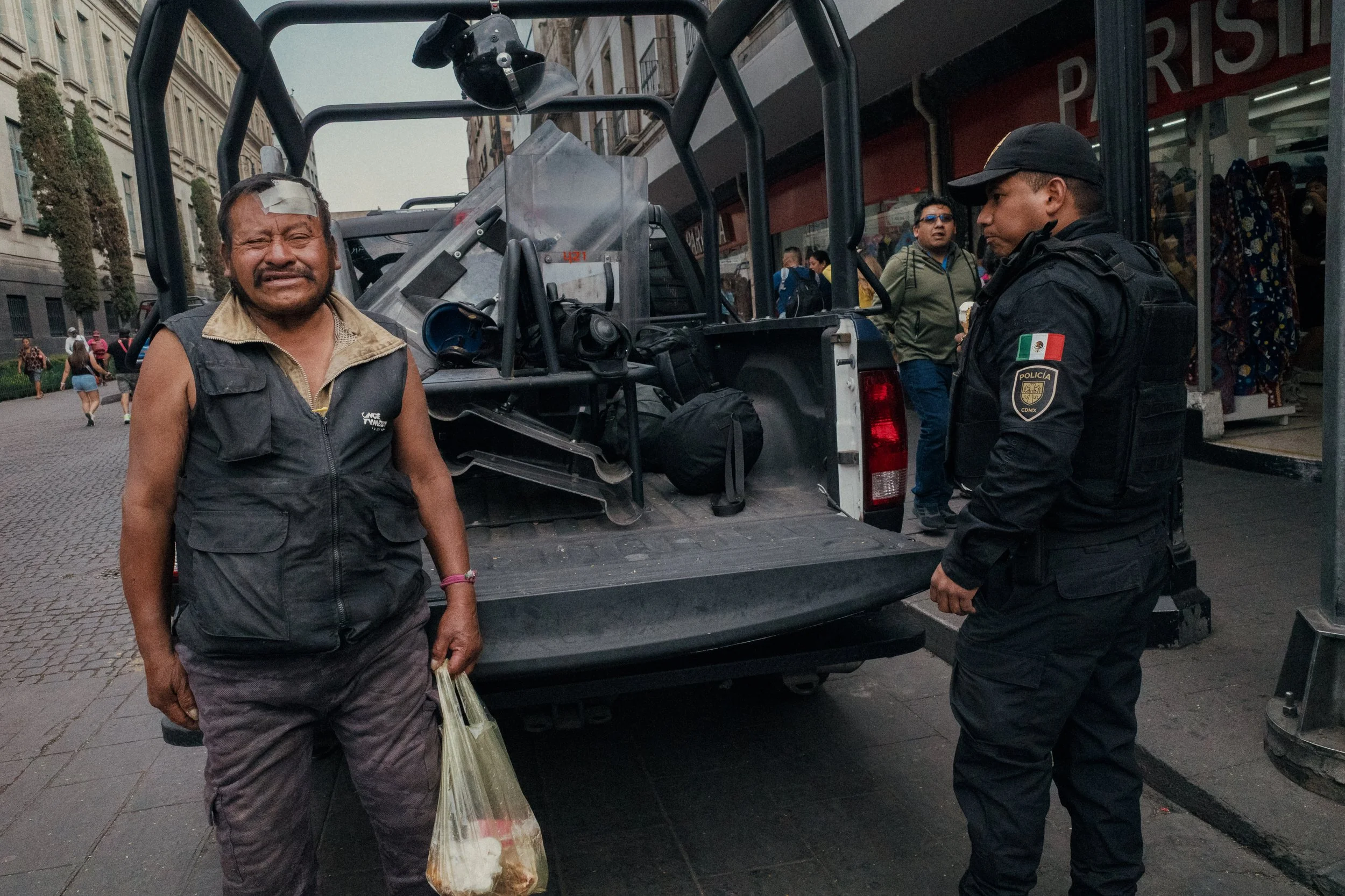 A man with a hurt forehead and a bandage, standing beside a police officer, in front of a police truck with equipment, on a city street. Several pedestrians are visible in the background.