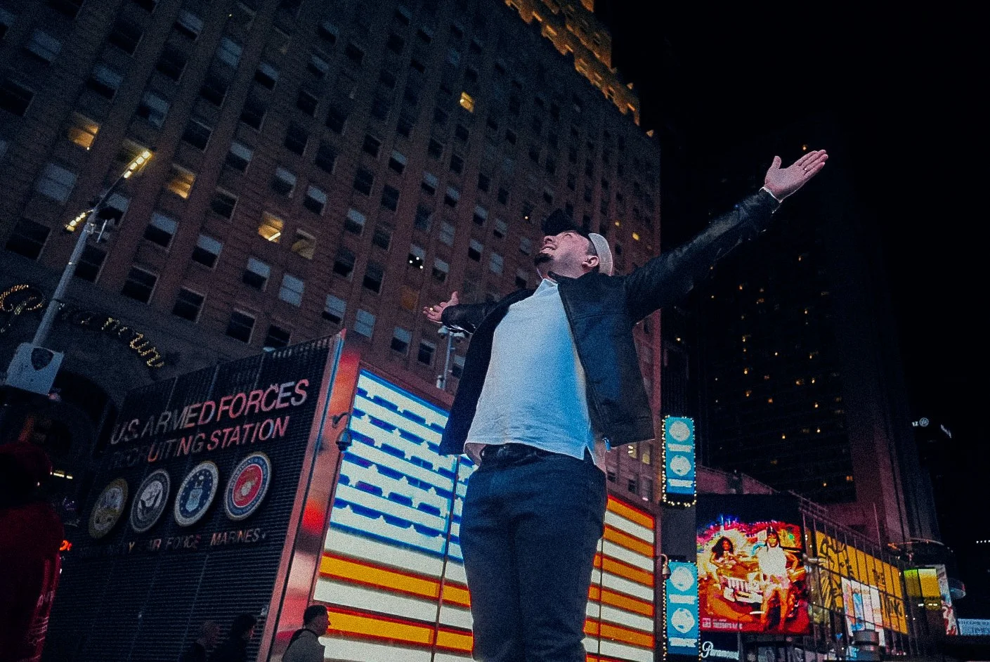 A man wearing a gray cap, white shirt, and black jacket stands with arms outstretched in Times Square at night, illuminated by digital billboards and neon lights.