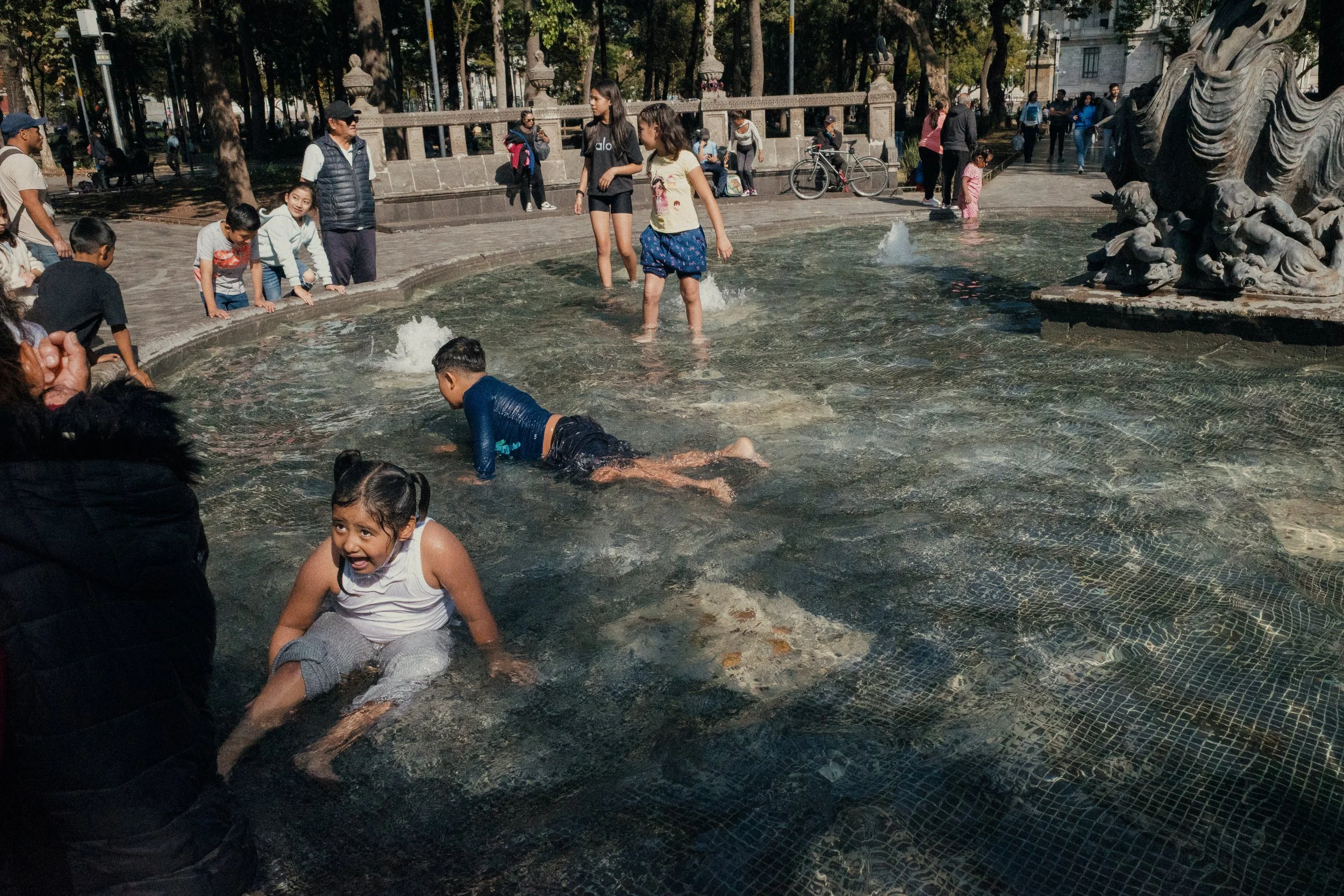 Children playing and splashing in a fountain in a park surrounded by onlookers and trees.